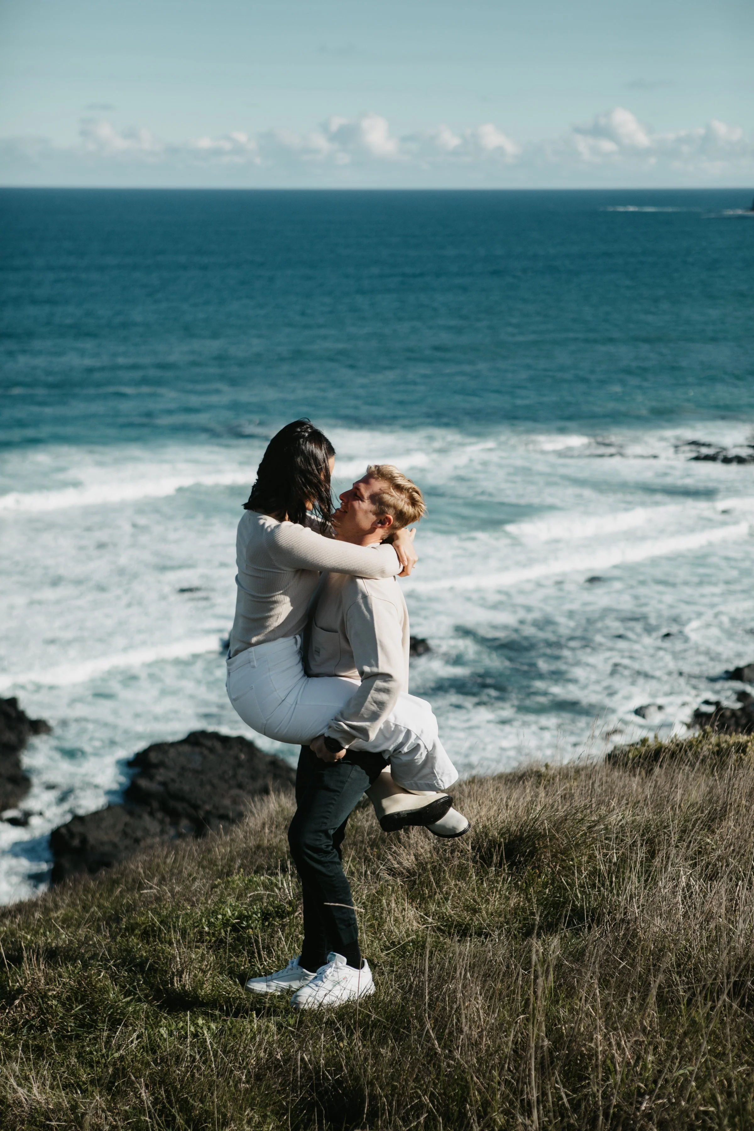 A couple is on a rocky coastline, with the man carrying the woman in his arms. They are embracing each other affectionately, with the ocean and a cloudy sky in the background.