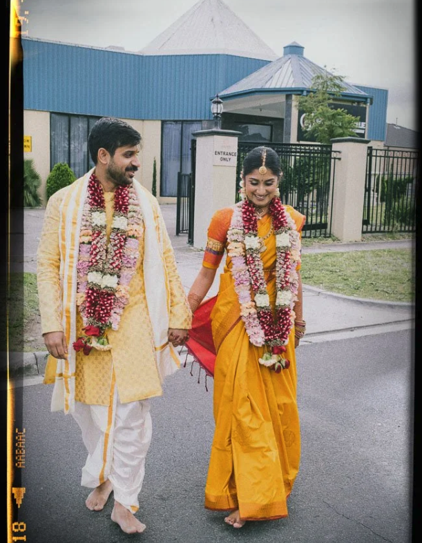 A couple dressed in traditional Indian attire, walking hand in hand outdoors, adorned with flower garlands, with a building and fence in the background.