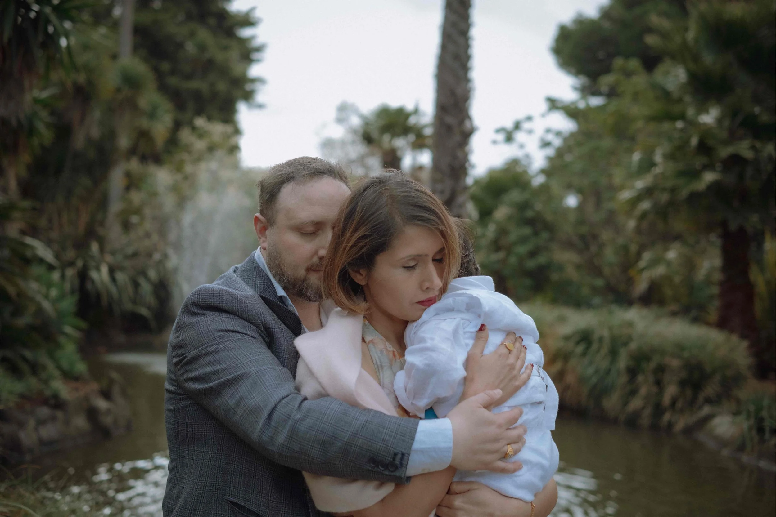 A man and woman holding a young girl close outdoors near a river, surrounded by trees.