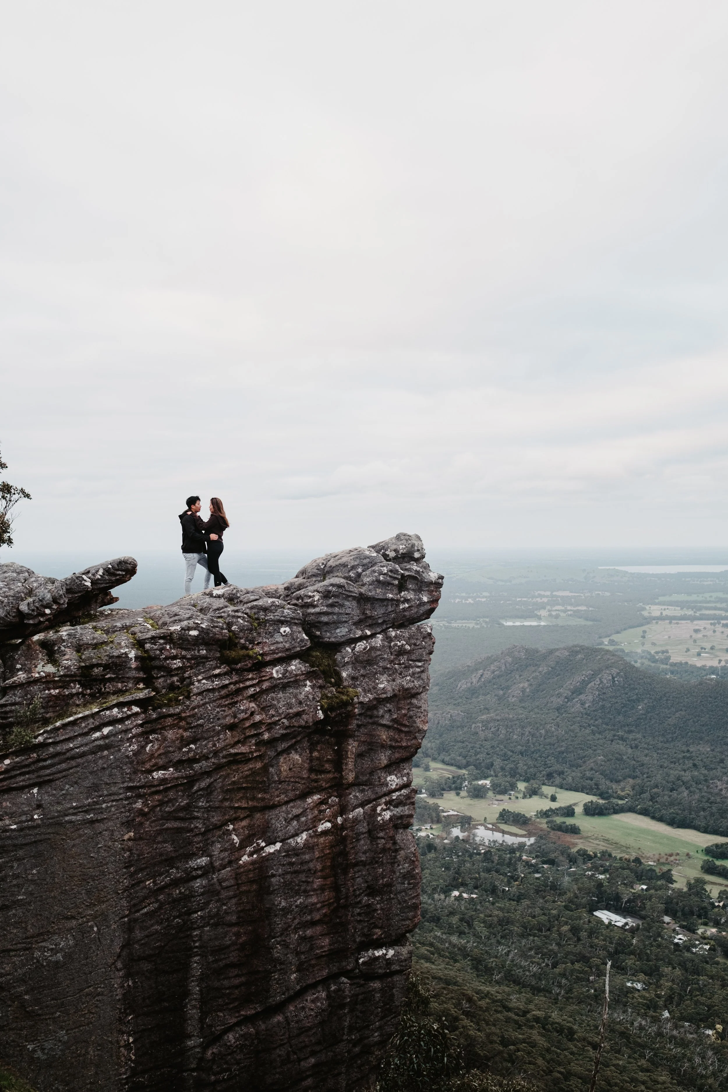 Two people standing on a rocky ledge on a mountain, with a scenic view of lush green valleys, trees, and distant hills under a cloudy sky.
