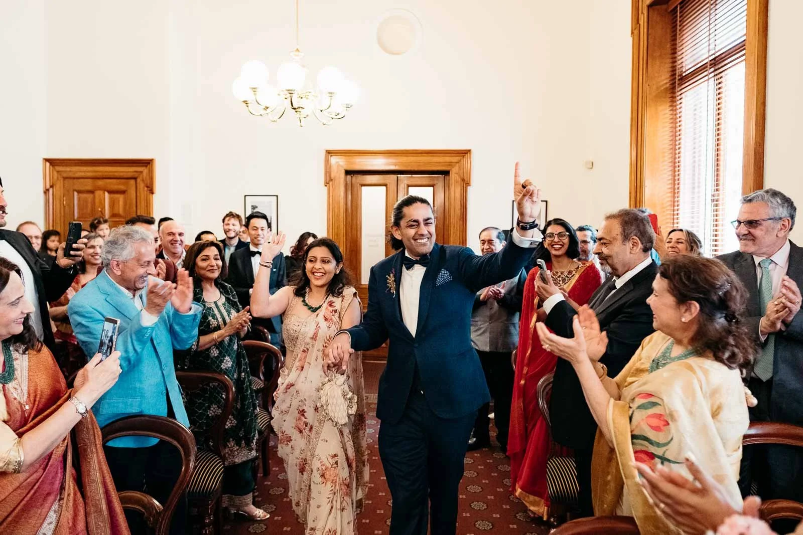 INDIAN WEDDING AT OLD TREASURY BUILDING WITH COUPLES WALKING TOGETHER DOWN THE ISLE AND WEDDING GUESTS CHEERING FOR THEM