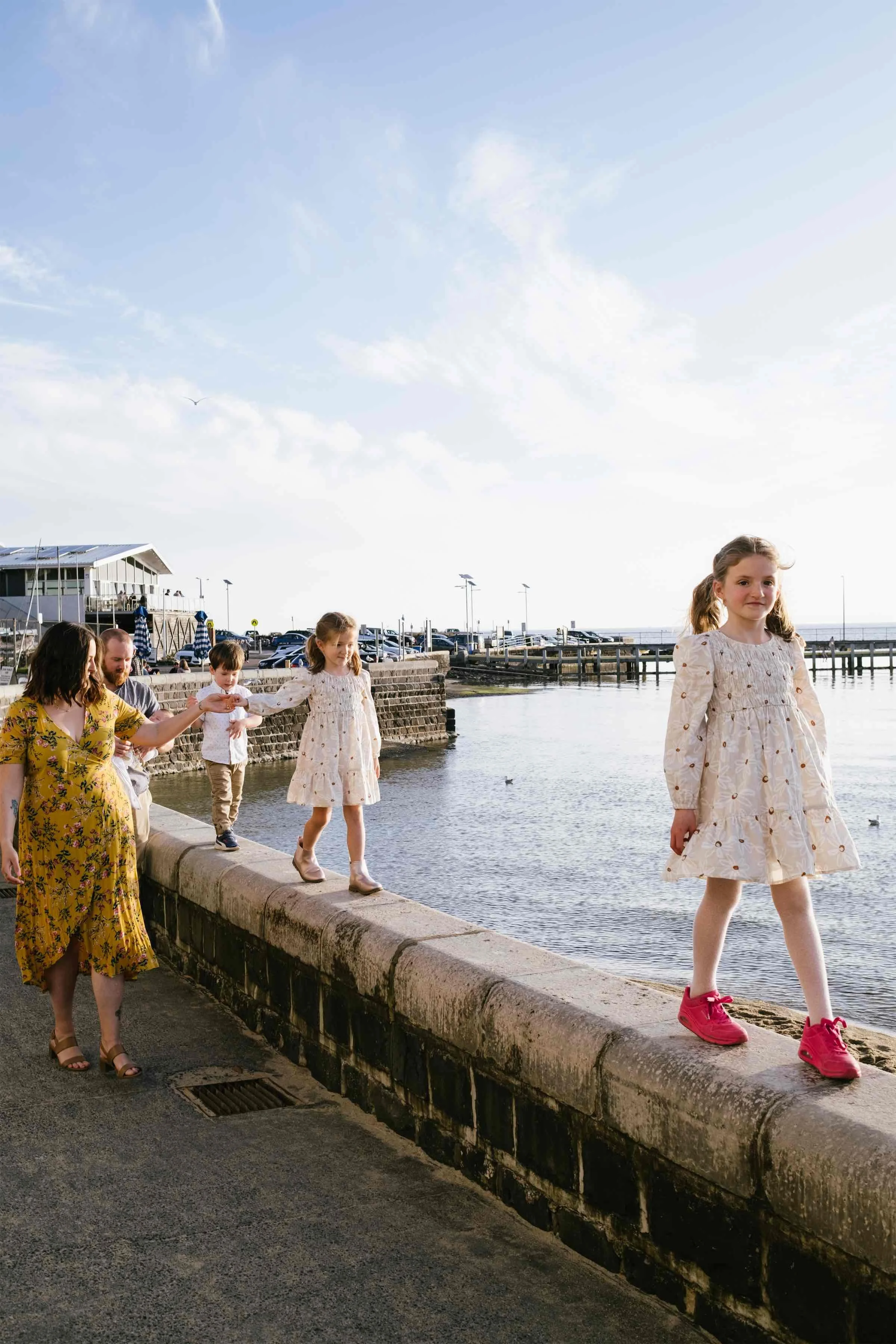 A group of people, including children and an adult woman, walking along a waterfront promenade during daytime. There is a girl in a cream-colored dress with red shoes standing on the edge of a stone wall, looking at the camera. Others are walking nea