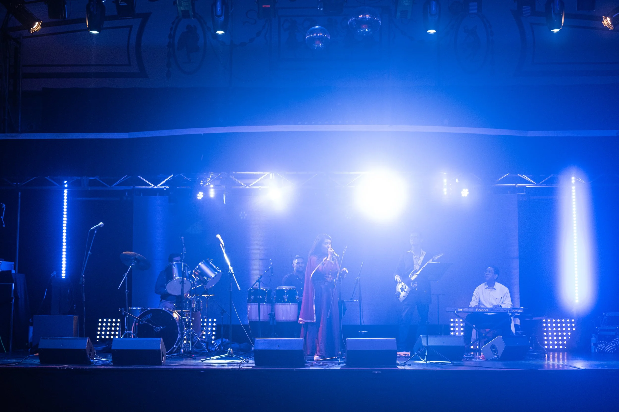 A live stage performance with a musician singing into a microphone, surrounded by band members playing drums, keyboard, and guitar under blue stage lighting and ceiling spotlights.