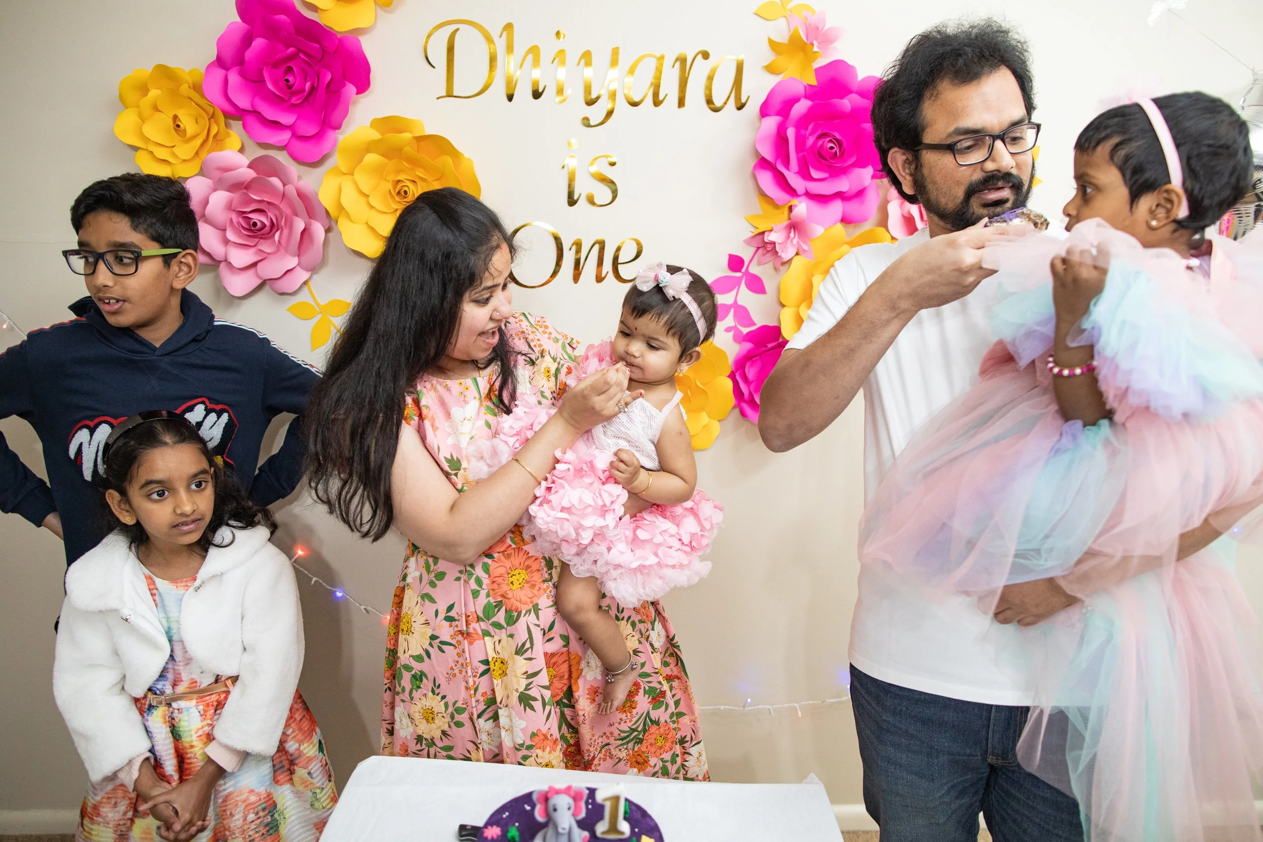 Children and adults at a birthday party, with a colorful paper flower backdrop and a birthday cake celebrating a first birthday.