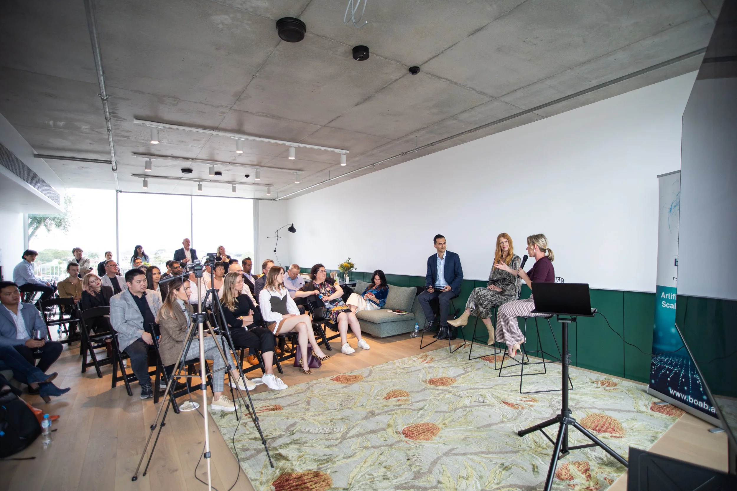 A large conference room filled with an audience watching a panel discussion. Three women and one man are seated on stage, with one woman speaking into a microphone. The room has large windows, wood flooring, and a carpeted area, with a mix of chairs 