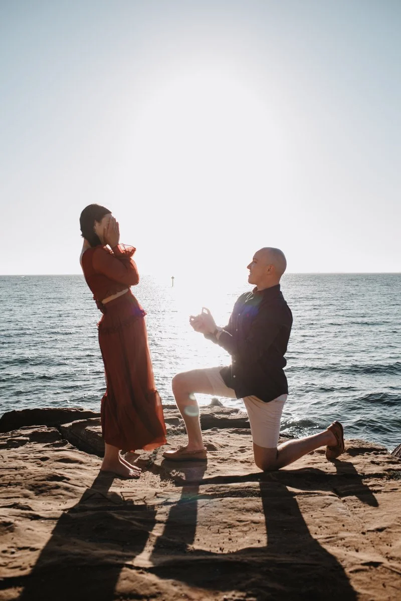 A man proposing marriage to a woman on a rocky beach during sunset, with the ocean in the background.