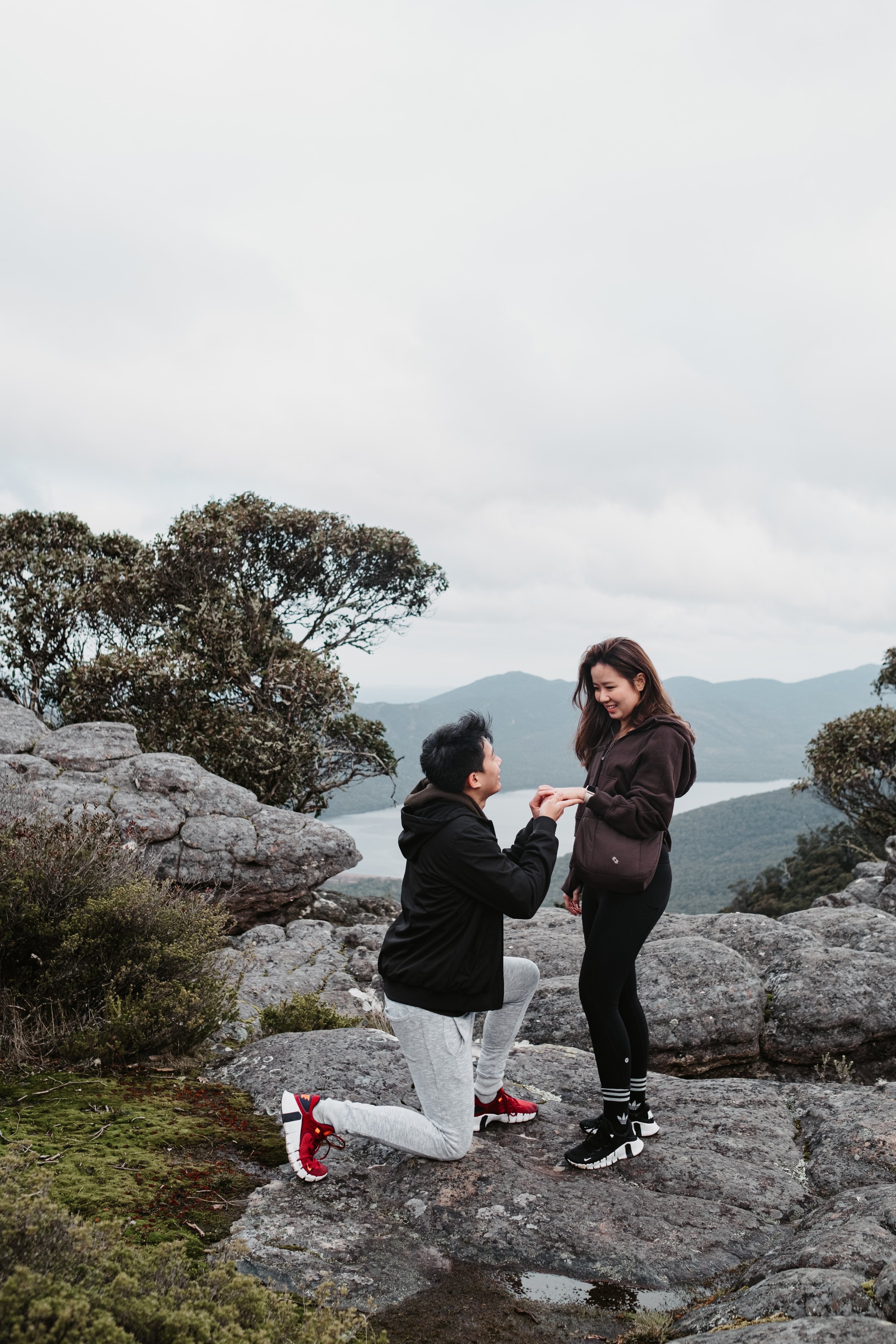 A man proposing marriage to a woman on a scenic rocky outdoor location with mountains and cloudy sky in the background.