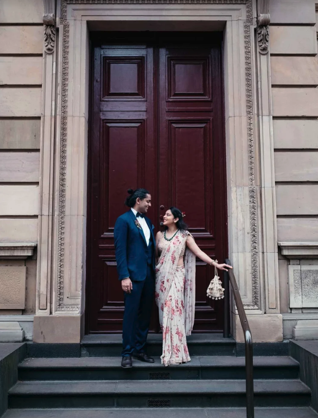 A man and woman in traditional and formal attire standing on steps in front of large wooden doors, smiling and looking at each other.