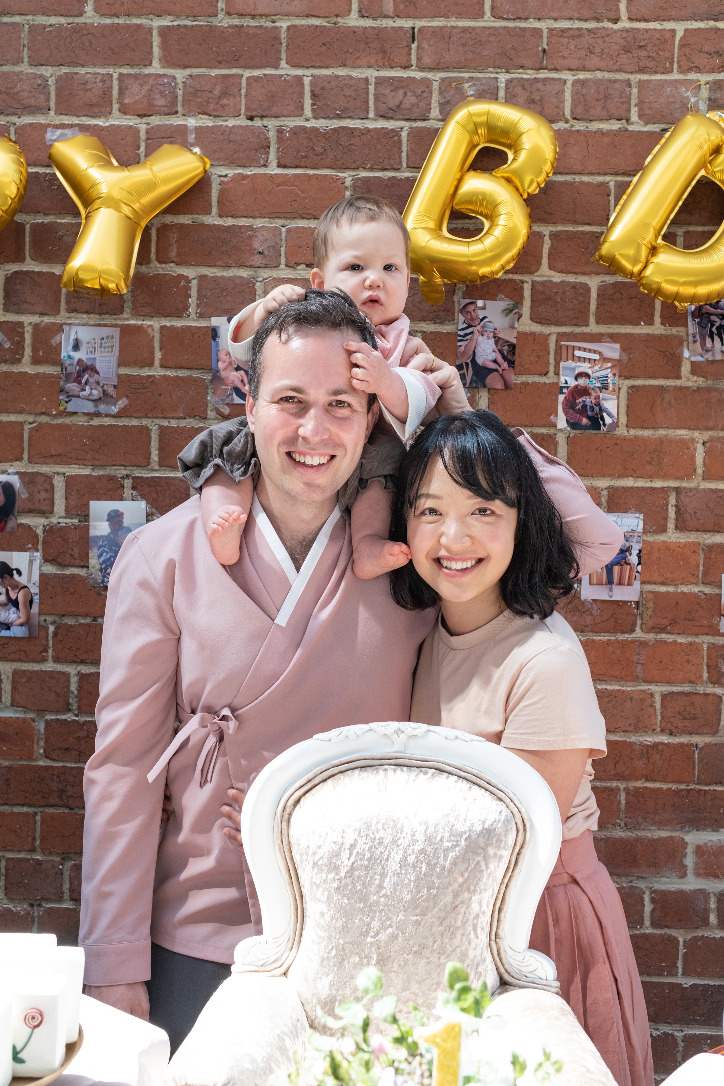 A family of three celebrating a birthday at a table in front of a brick wall. The man is wearing a pink traditional outfit, the woman is in a beige shirt, and the child, sitting on the man's shoulders, is reaching out. There are gold balloons that sp