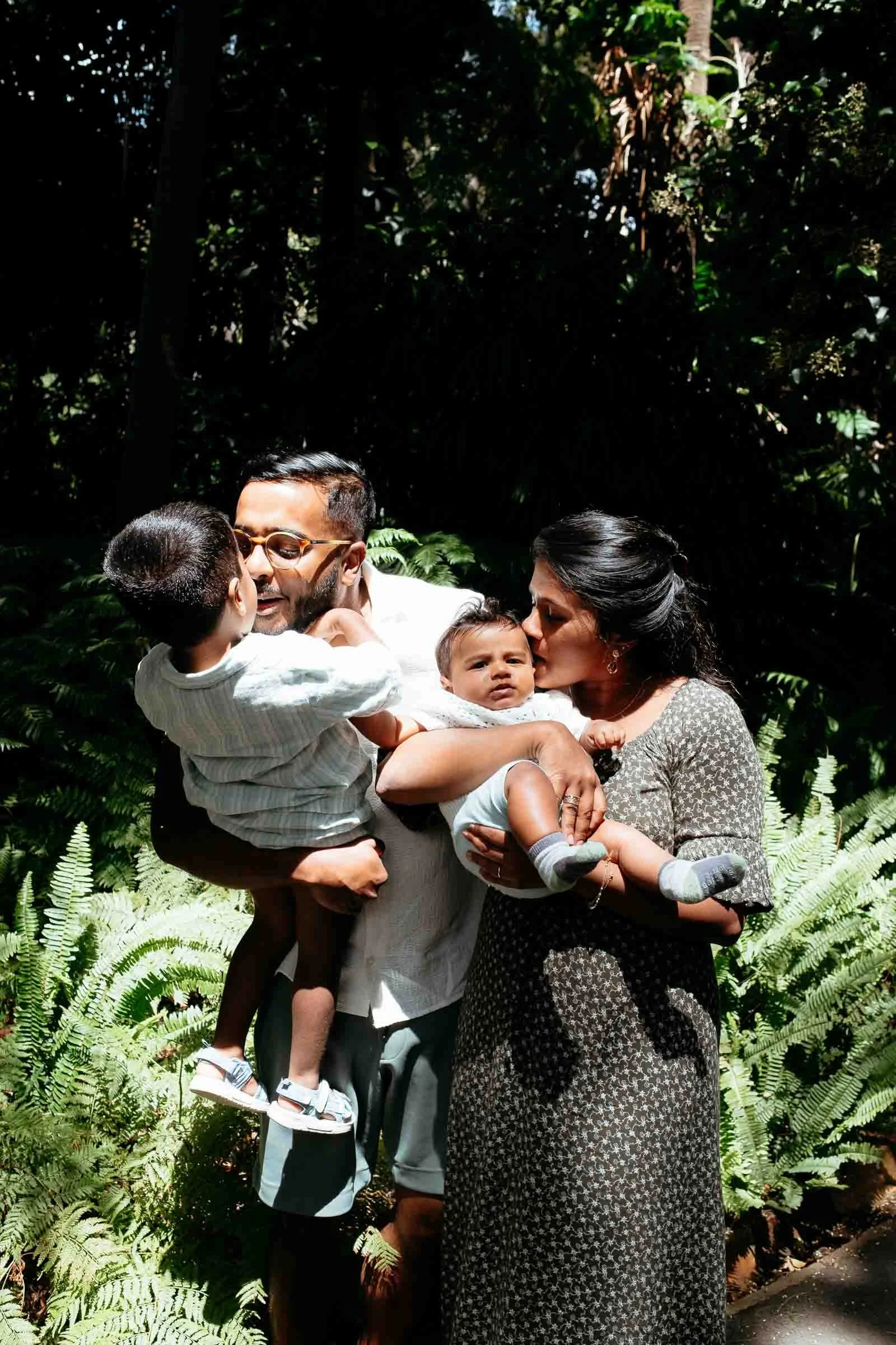 A family of four outdoors among lush green foliage, including two children and two adults, with the adults holding and kissing the children.