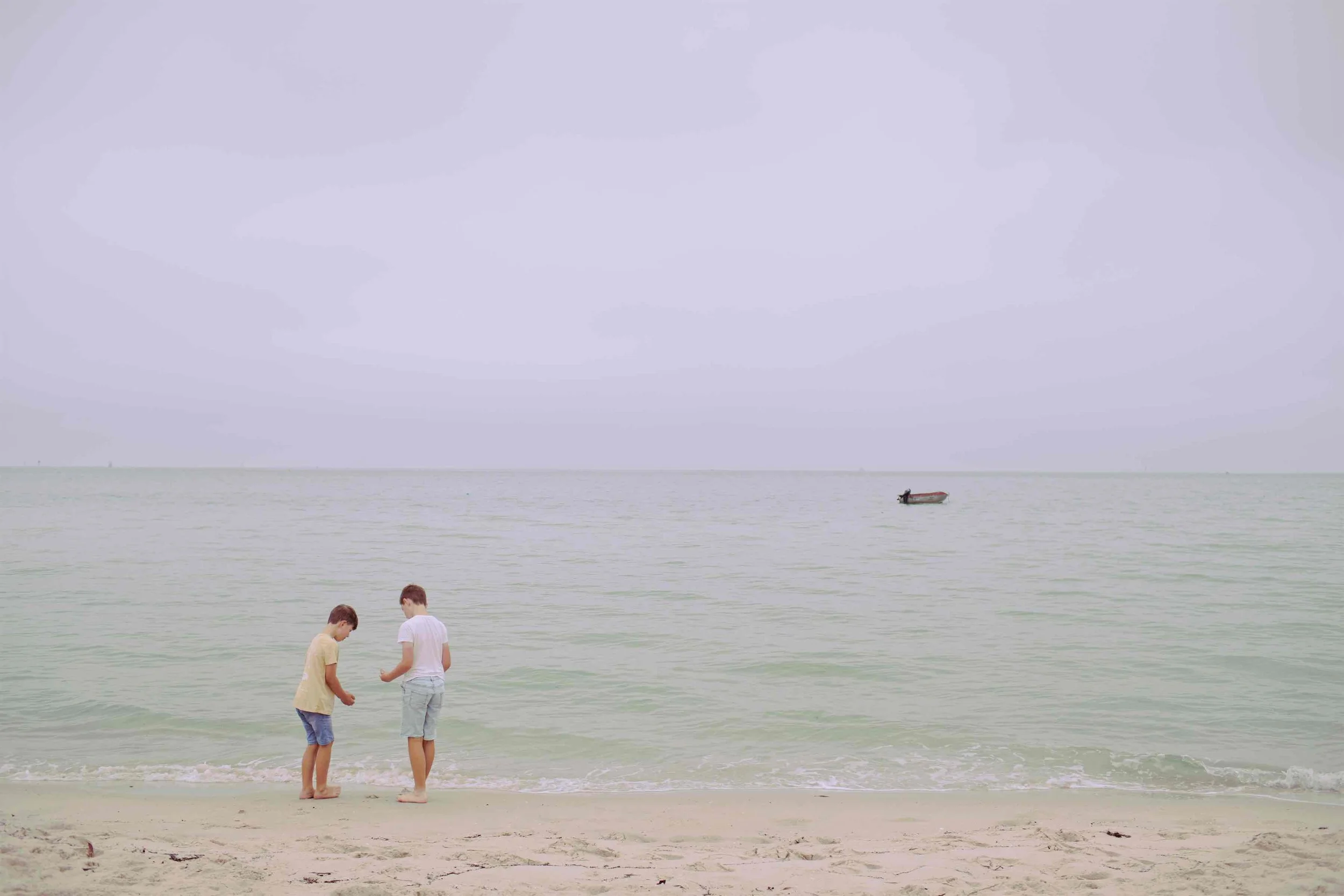 Three children standing at the shoreline on a beach, looking down at the sand and water with a small boat floating in the distance on a calm sea, under a cloudy sky.