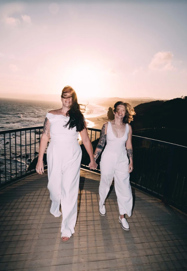 Two women in white jumpsuits holding hands walk on a wooden bridge near the ocean with the sun setting behind them, casting a warm glow.