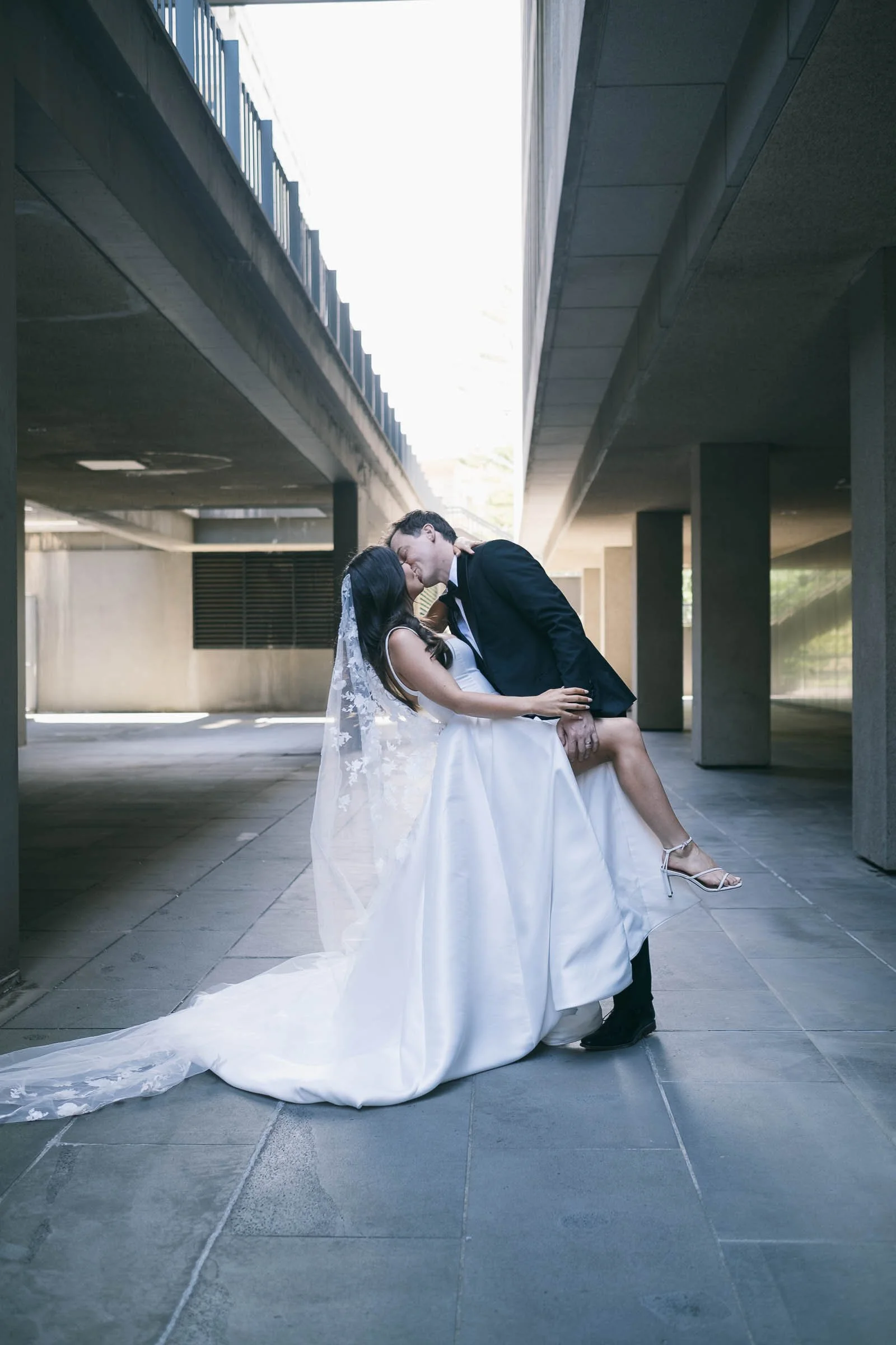 Bride and groom kissing under a modern architectural structure, bride in a white gown with a veil and groom in a black suit, embracing romantically.