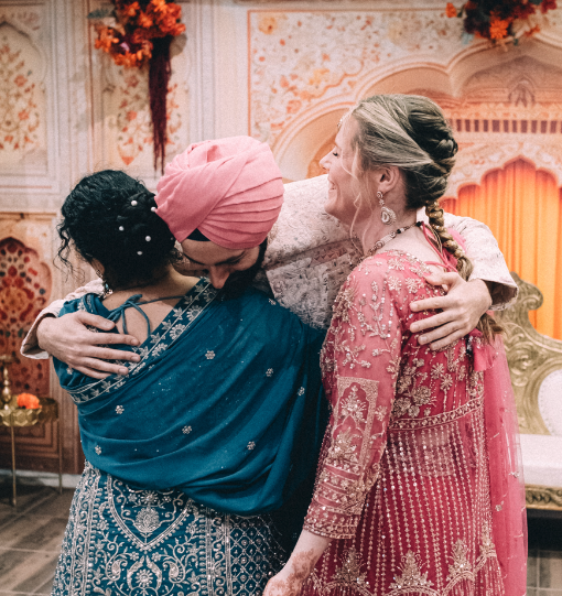 Three women hugging, dressed in traditional Indian and South Asian clothing at a festive event with ornate decorations and floral arrangements in the background.
