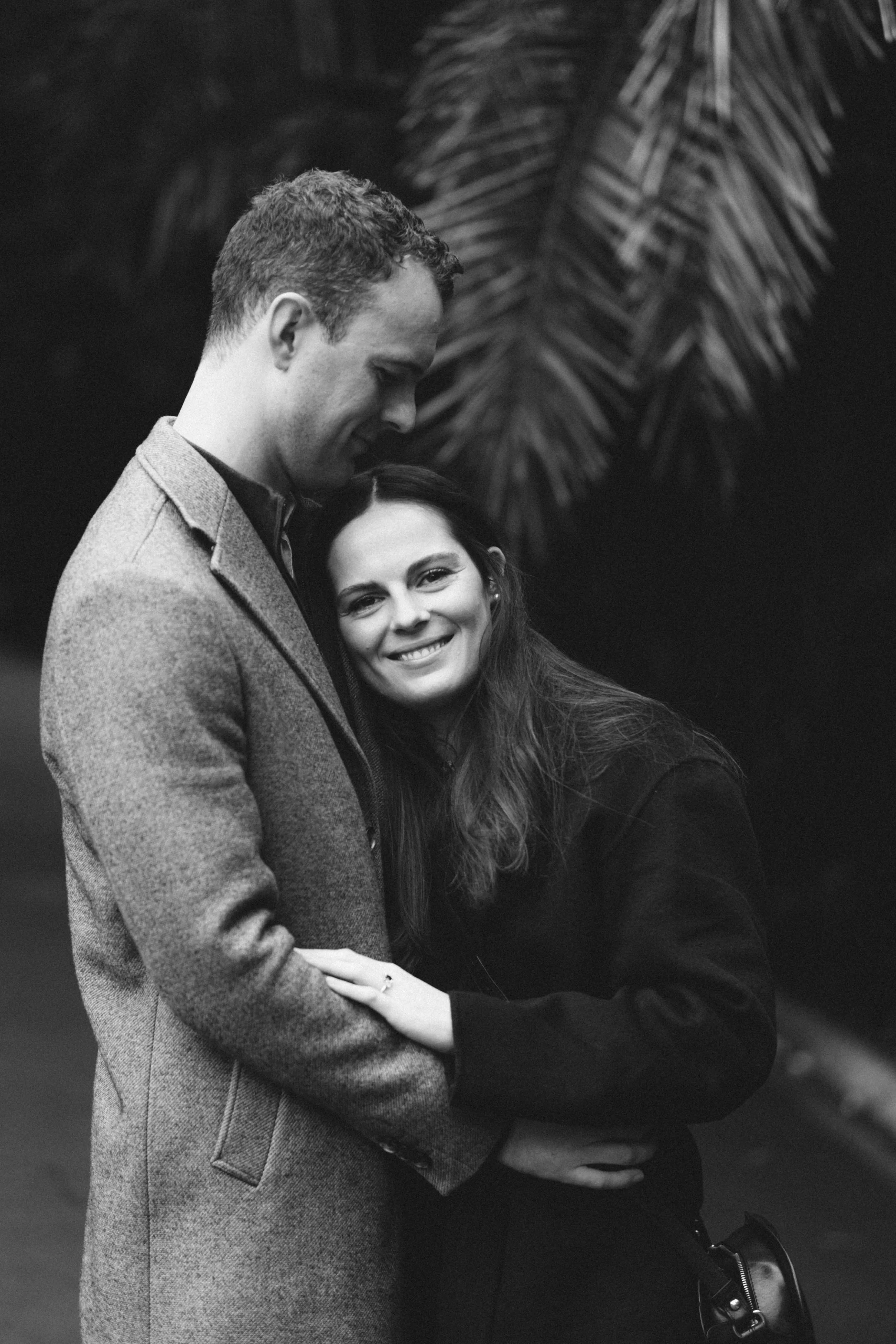 A black and white photo of a man and woman embracing, with the man smiling and resting his forehead on the woman's head, with large palm leaves in the background.