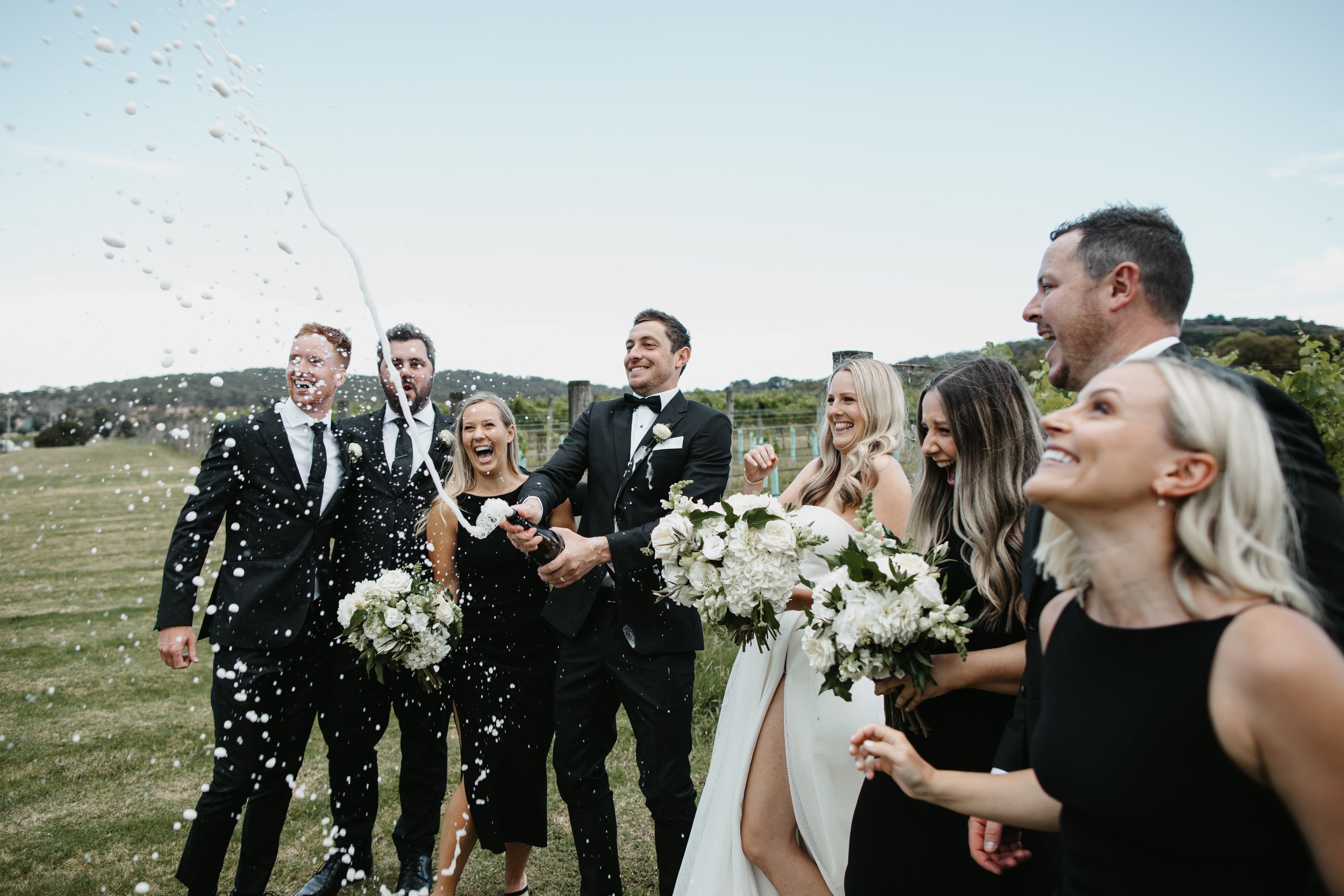 A wedding party joyously sprays champagne outdoors. The group is dressed in formal attire, with women holding white flower bouquets. The background features green fields and a clear sky.