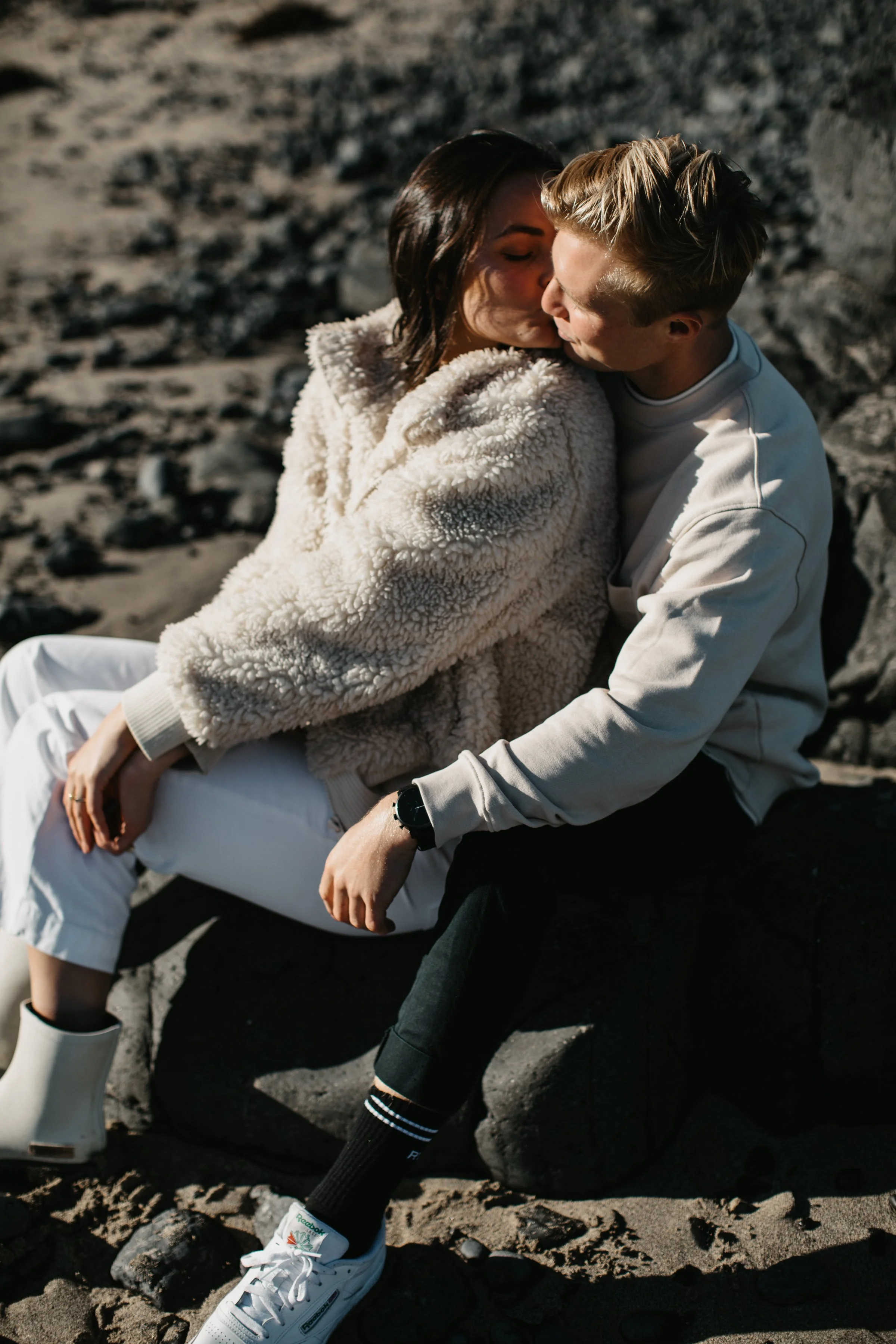 A young couple sitting on a rocky beach, sharing an intimate kiss. The woman has dark hair and is wearing a fluffy beige jacket and white pants. The man has light hair and is dressed in a white sweatshirt and black pants. They are surrounded by volca