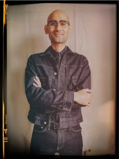 A man with glasses wearing a black denim jacket and black pants, standing with arms crossed in front of a neutral background.