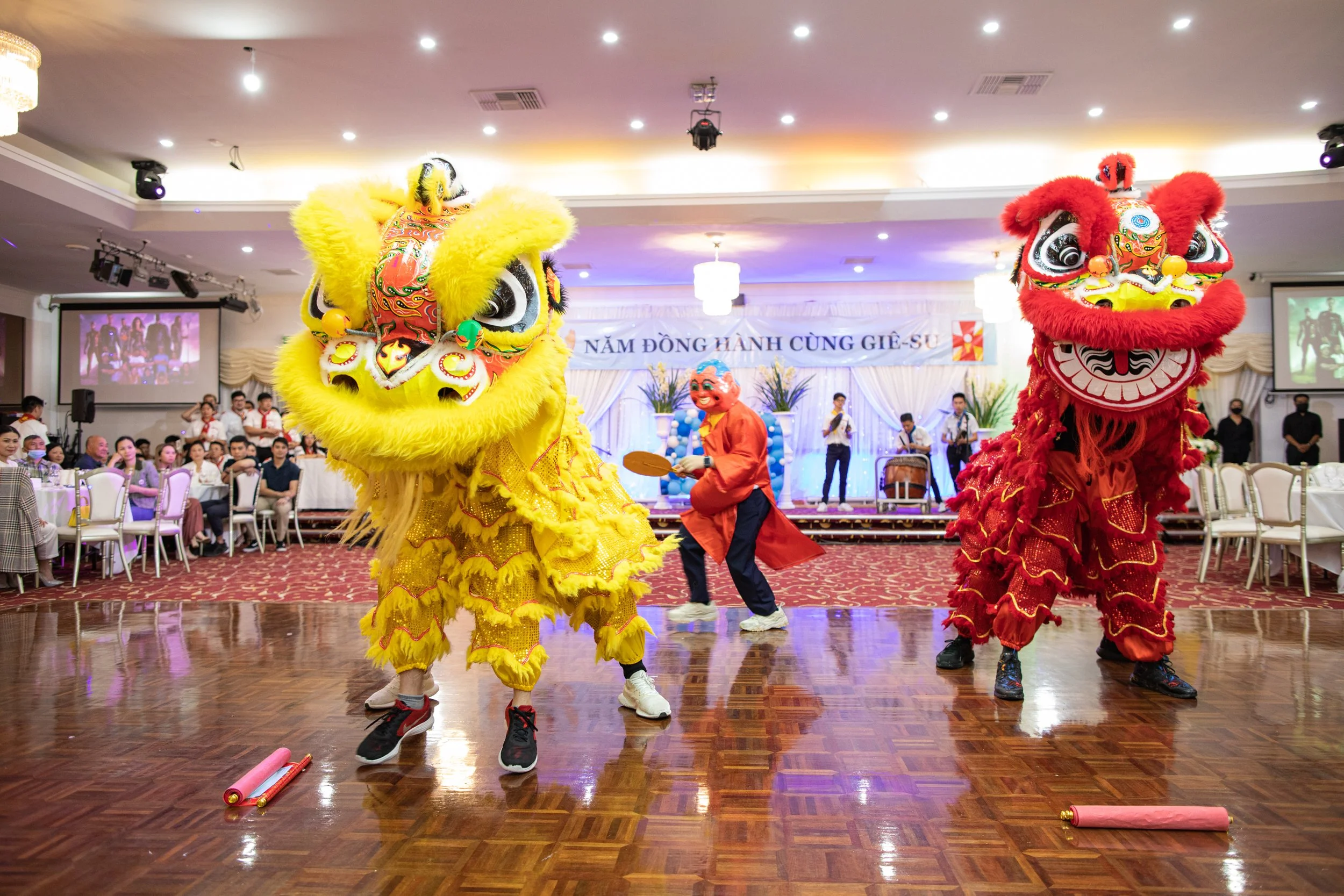 Lion dancers performing at an indoor celebration, with a crowd watching. The dancers wear colorful costumes with large lion heads, one yellow and one red, and are dancing on a wooden floor. There are plates of rolled paper on the floor in front of th