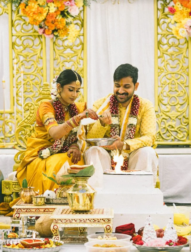 Indian bride and groom during wedding ceremony, seated on stage with decorations, floral arrangements, and traditional offerings.
