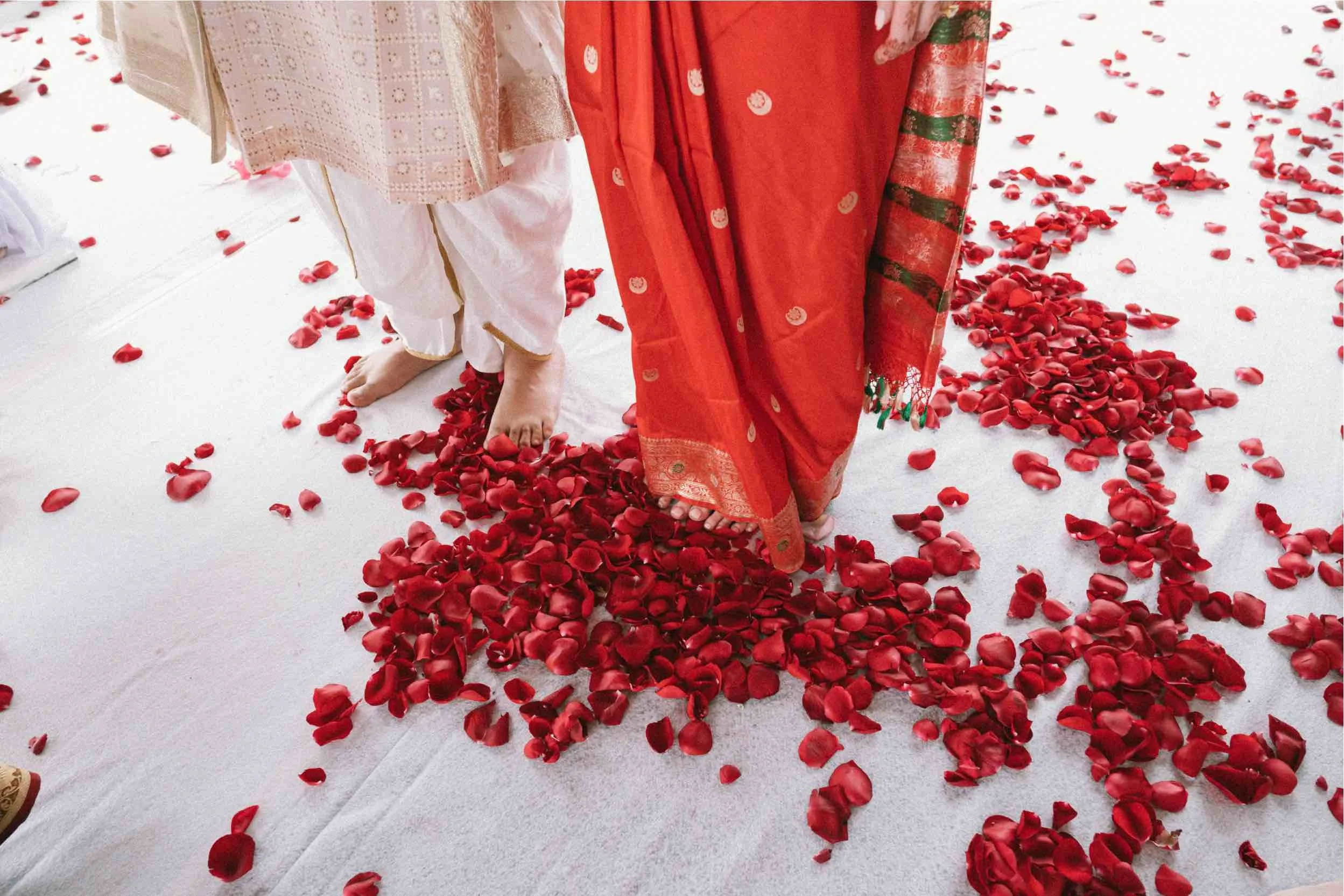 Close-up of women's feet standing on red rose petals scattered on a white surface, with women wearing traditional colorful dresses.