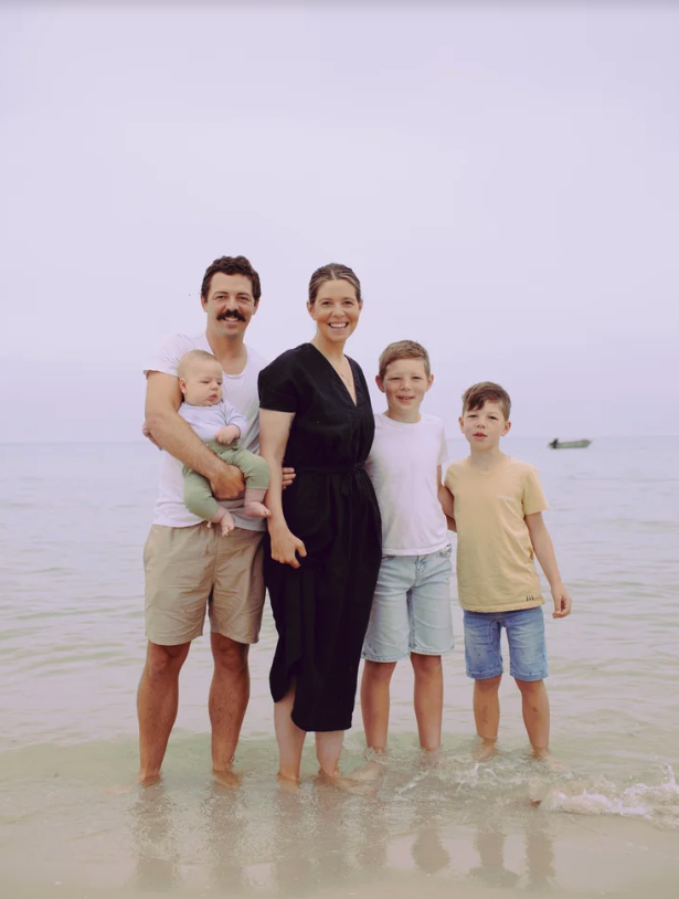 A family of five standing in shallow ocean water at the beach, smiling at the camera. The father is holding a baby, the mother is standing in the center, with two boys on her right.