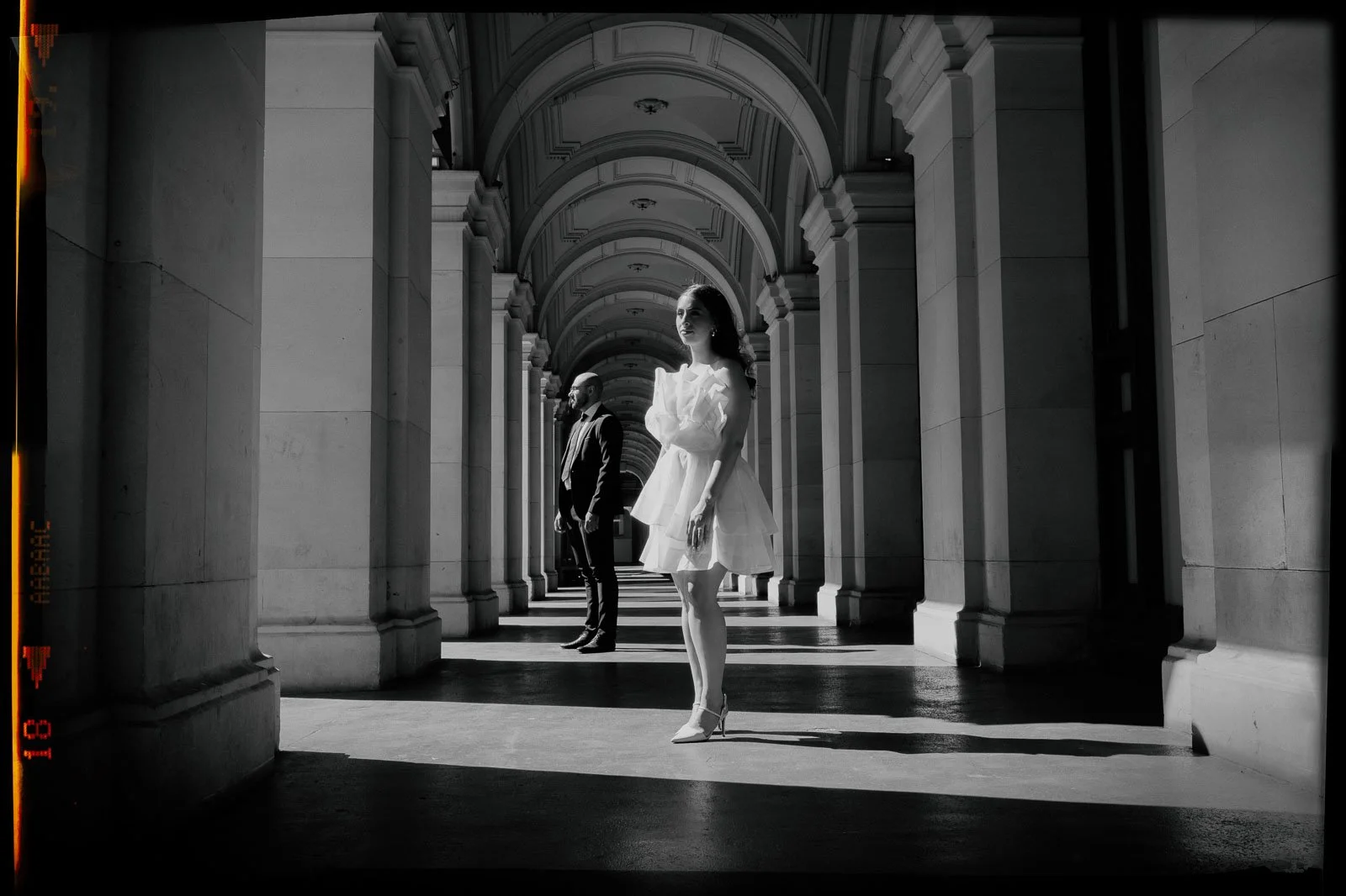Black and white photo of a woman in a white dress and high heels standing in a colonnade, with a man in a suit in the background