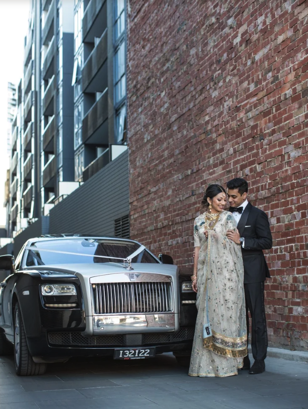 A couple dressed in formal wedding attire, standing next to a luxury car, against a brick wall in an urban setting.