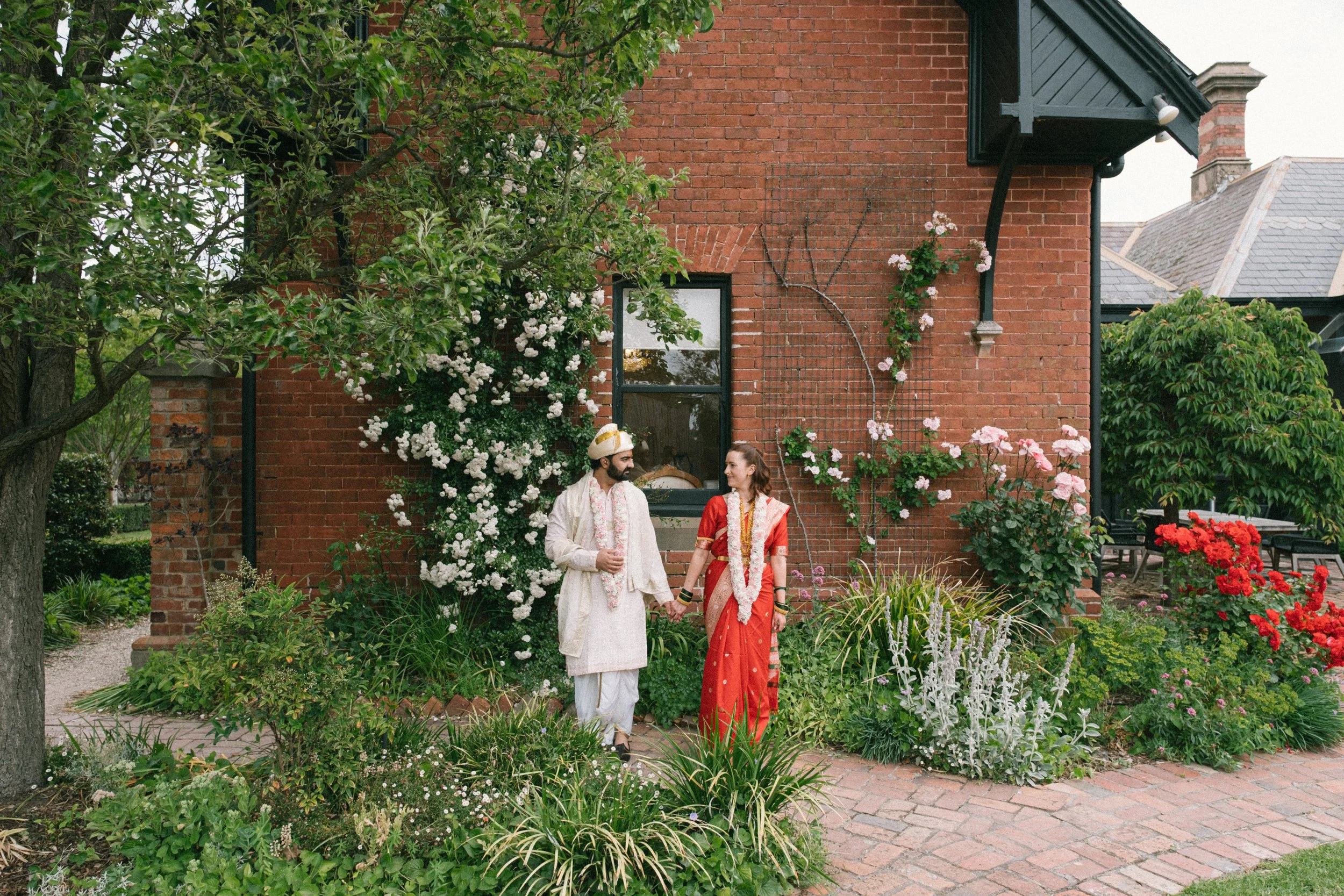 A couple dressed in traditional Indian attire holding hands in a garden with colorful flowers, trees, and a brick house in the background.