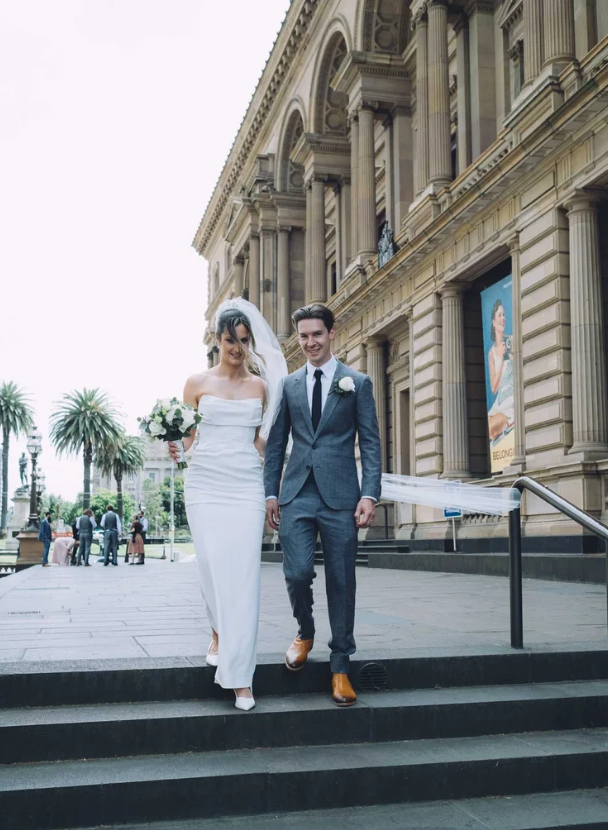 A bride and groom walking down steps outside a historic building, with the bride in a white strapless wedding gown holding a bouquet, and the groom in a gray suit with brown shoes, both smiling. Old Treasury Building Wedding