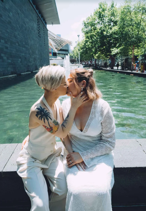 Two women kiss by a fountain in an outdoor urban setting with trees and modern buildings in the background.