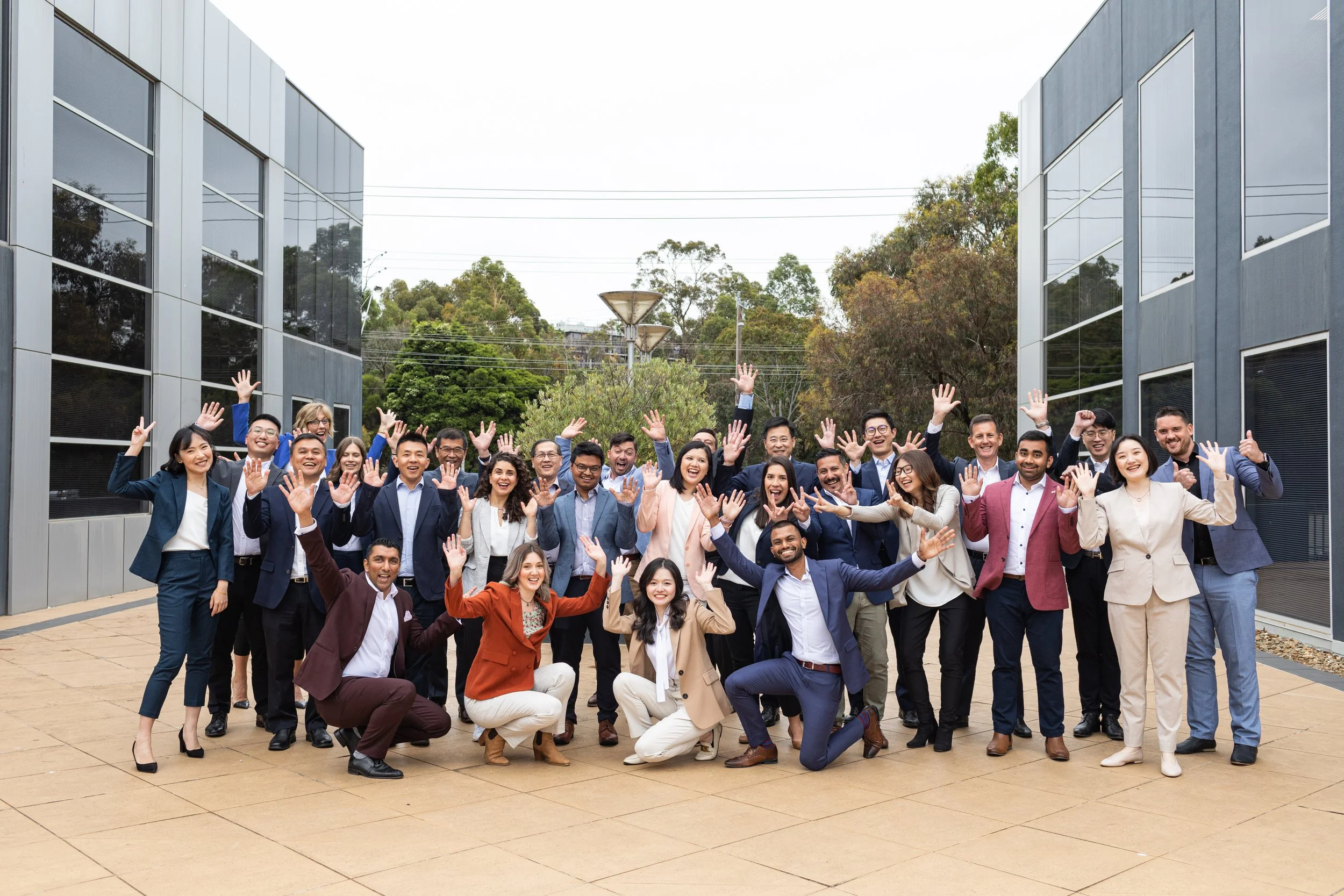 A group of diverse business people gathered outdoors, smiling and waving at the camera, with modern buildings and trees in the background.
