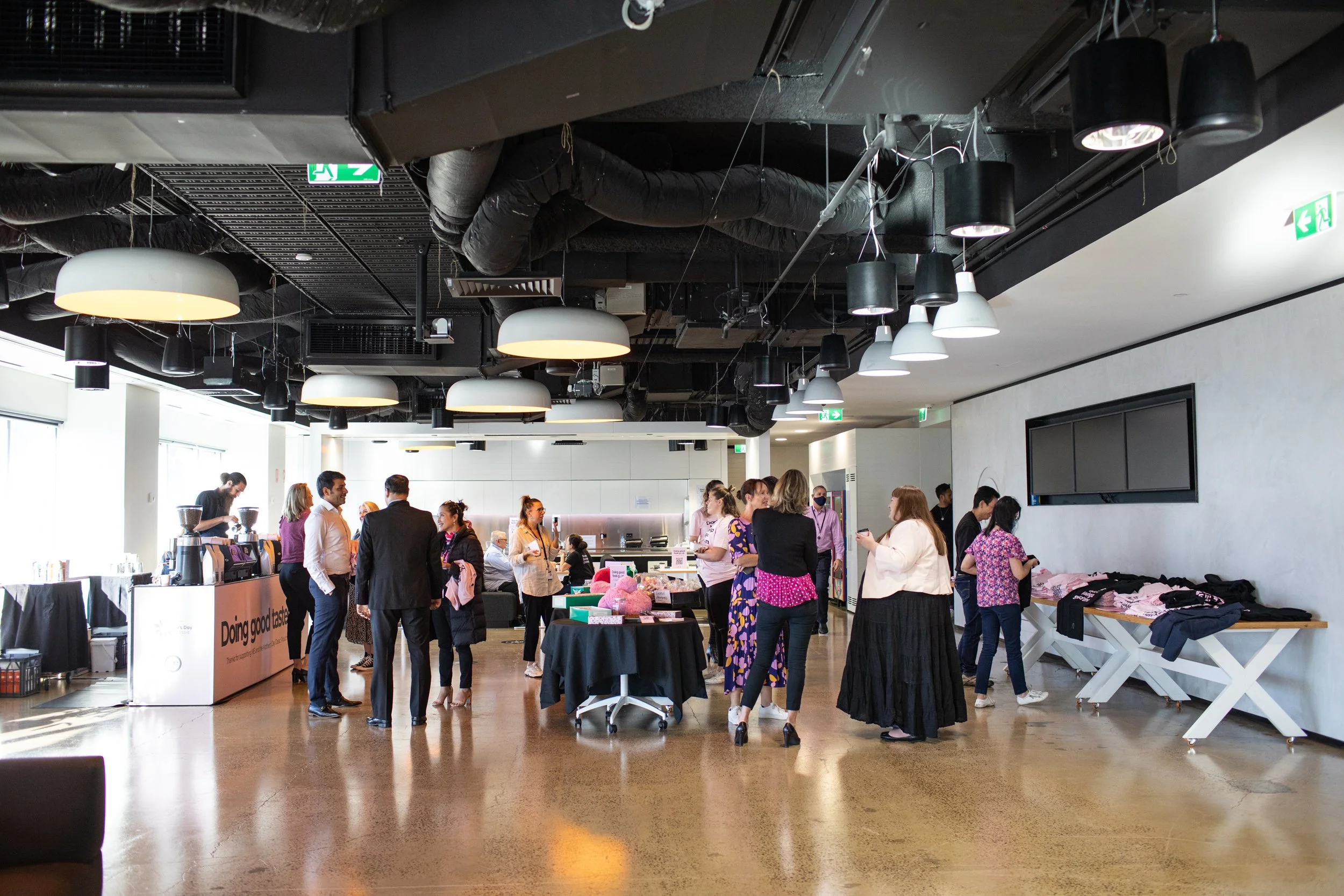 People gathered in an indoor event space, some are talking, others browsing tables with items for sale or display. The ceiling has exposed ducts and hanging light fixtures, and large windows let in natural light.