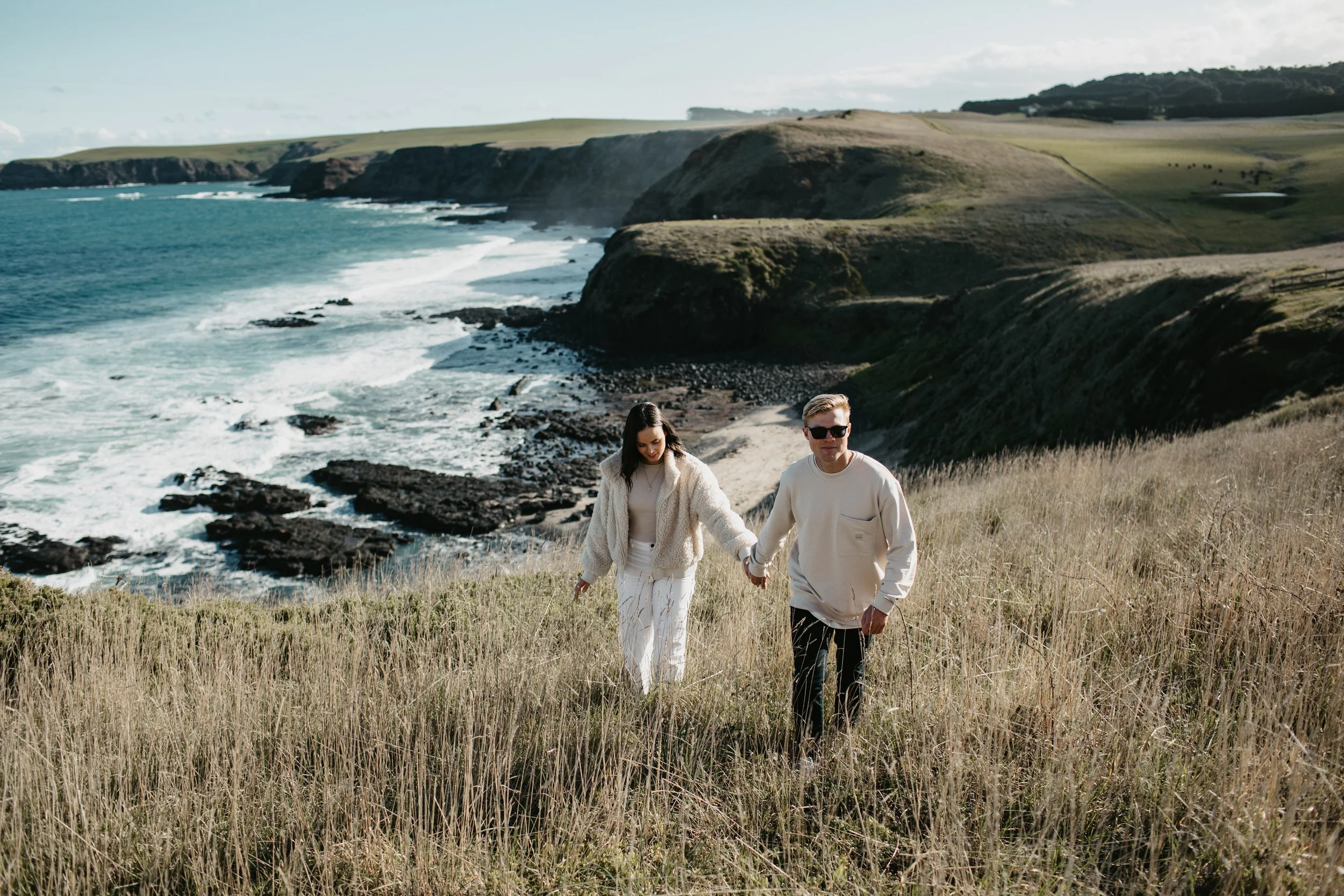 A couple holding hands walking through tall grass on a hillside overlooking the ocean with cliffs and waves, under a partly cloudy sky.