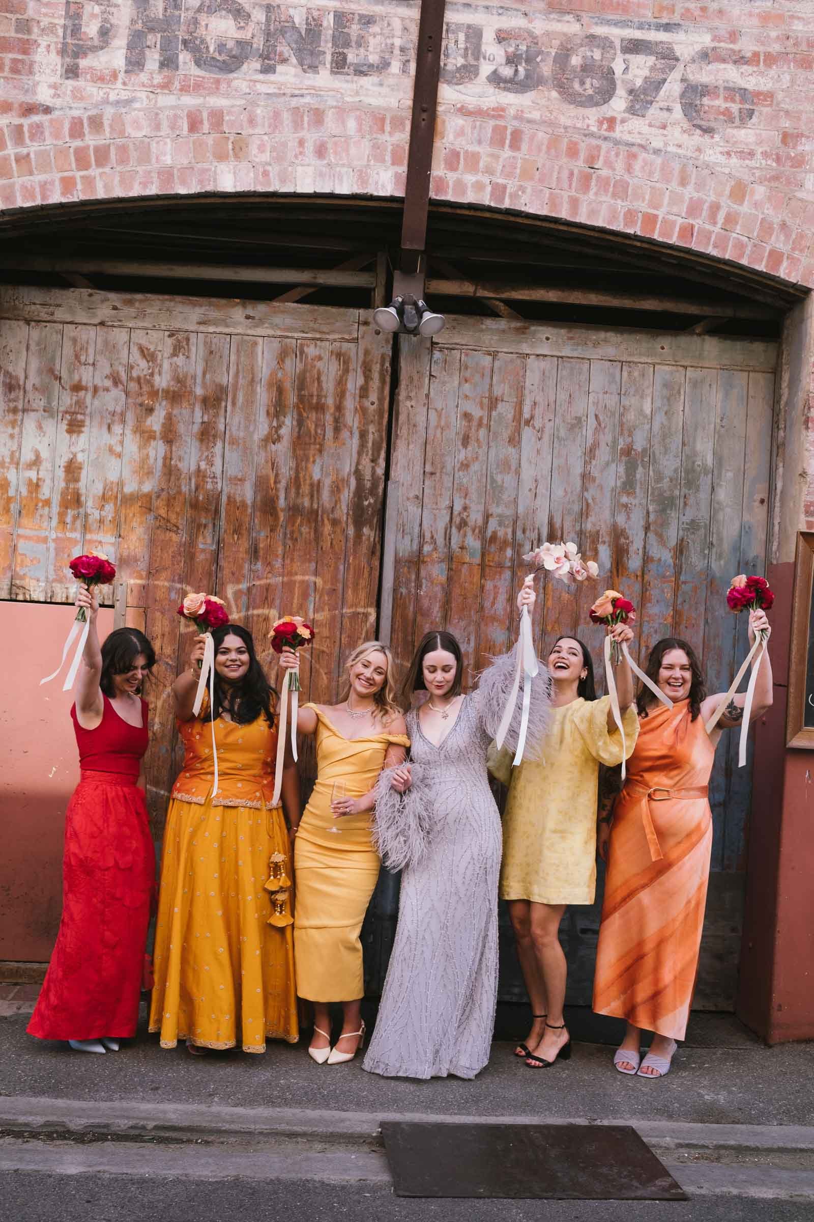 Group of women dressed in colorful dresses holding bouquets of flowers, standing in front of a rustic wooden gate with a brick wall background.