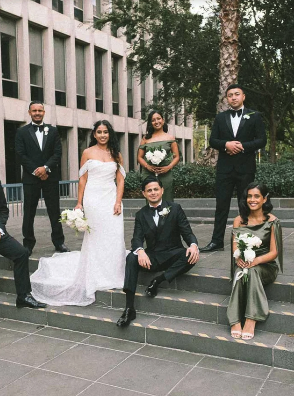 Group of seven young adults in formal attire, posing outdoors on steps in a city setting with a building and greenery in the background, celebrating a wedding.