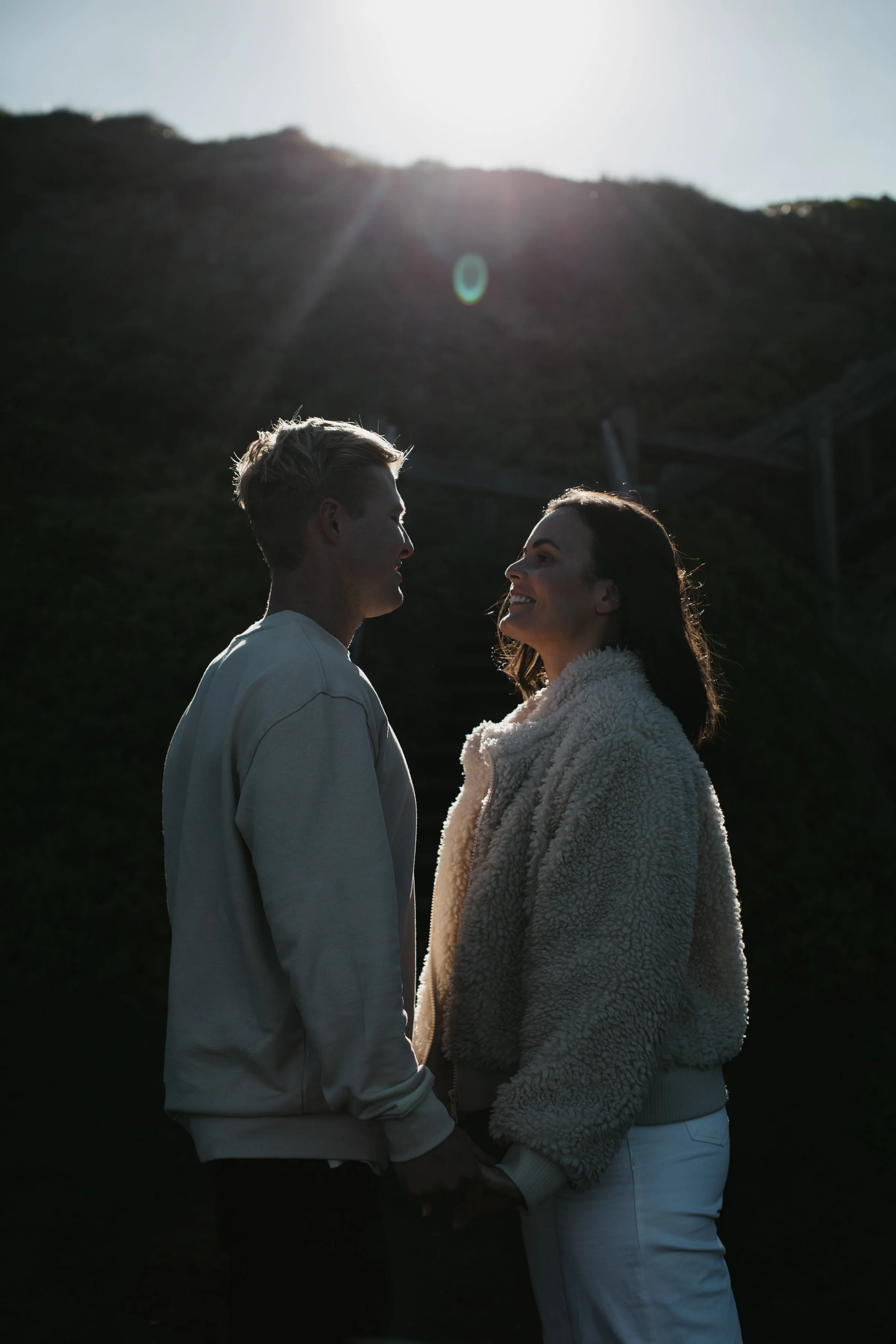 A man and a woman holding hands and facing each other outdoors, with a mountain and bright sun in the background.