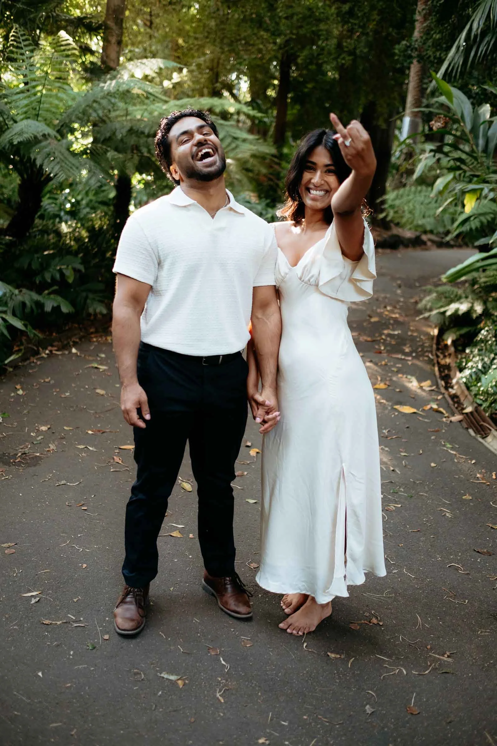 A couple, holding hands, standing outdoors in a lush green garden. The man is wearing a white polo shirt, black pants, and brown shoes, laughing with his head tilted back. The woman is wearing a white dress, barefoot, smiling, and showing her middle 