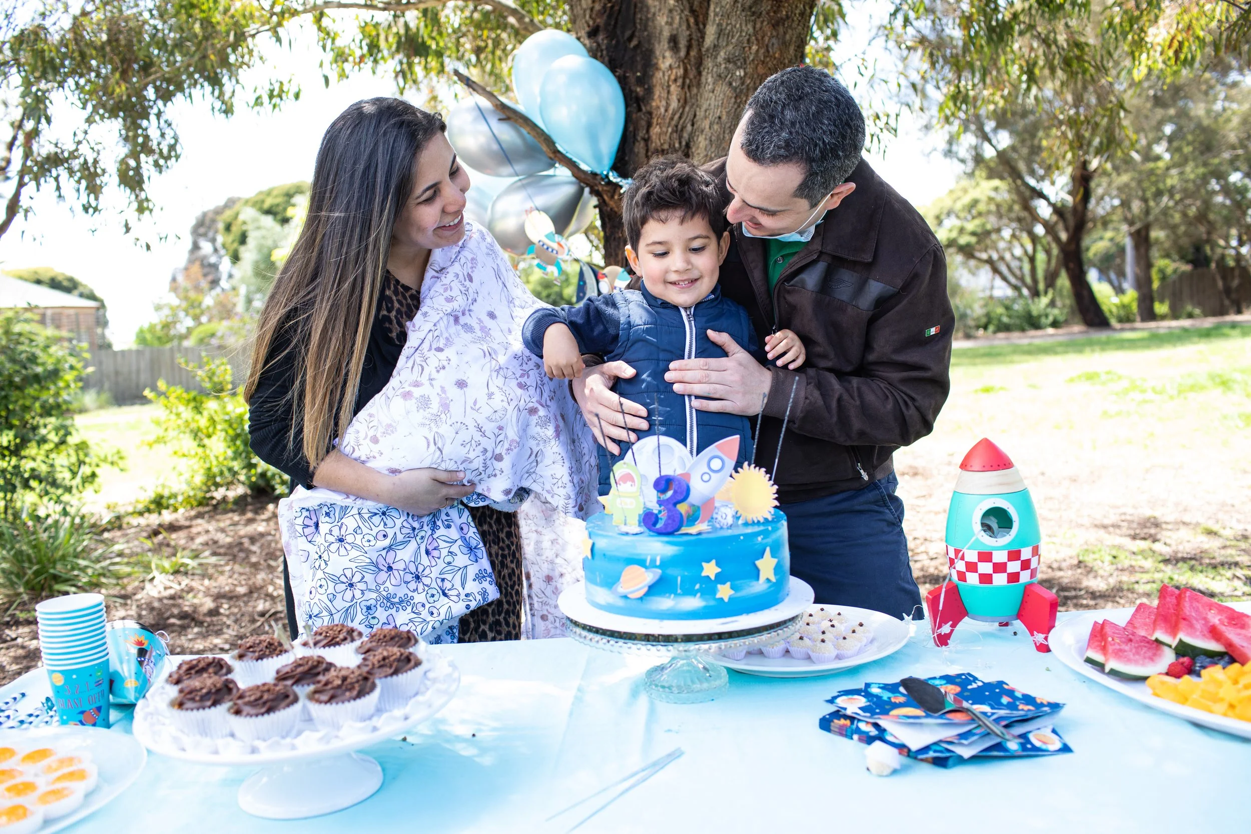 Family celebrating a child's birthday outdoors with a blue space-themed cake, balloons, and party decorations. The child, two adults, and party food are visible.