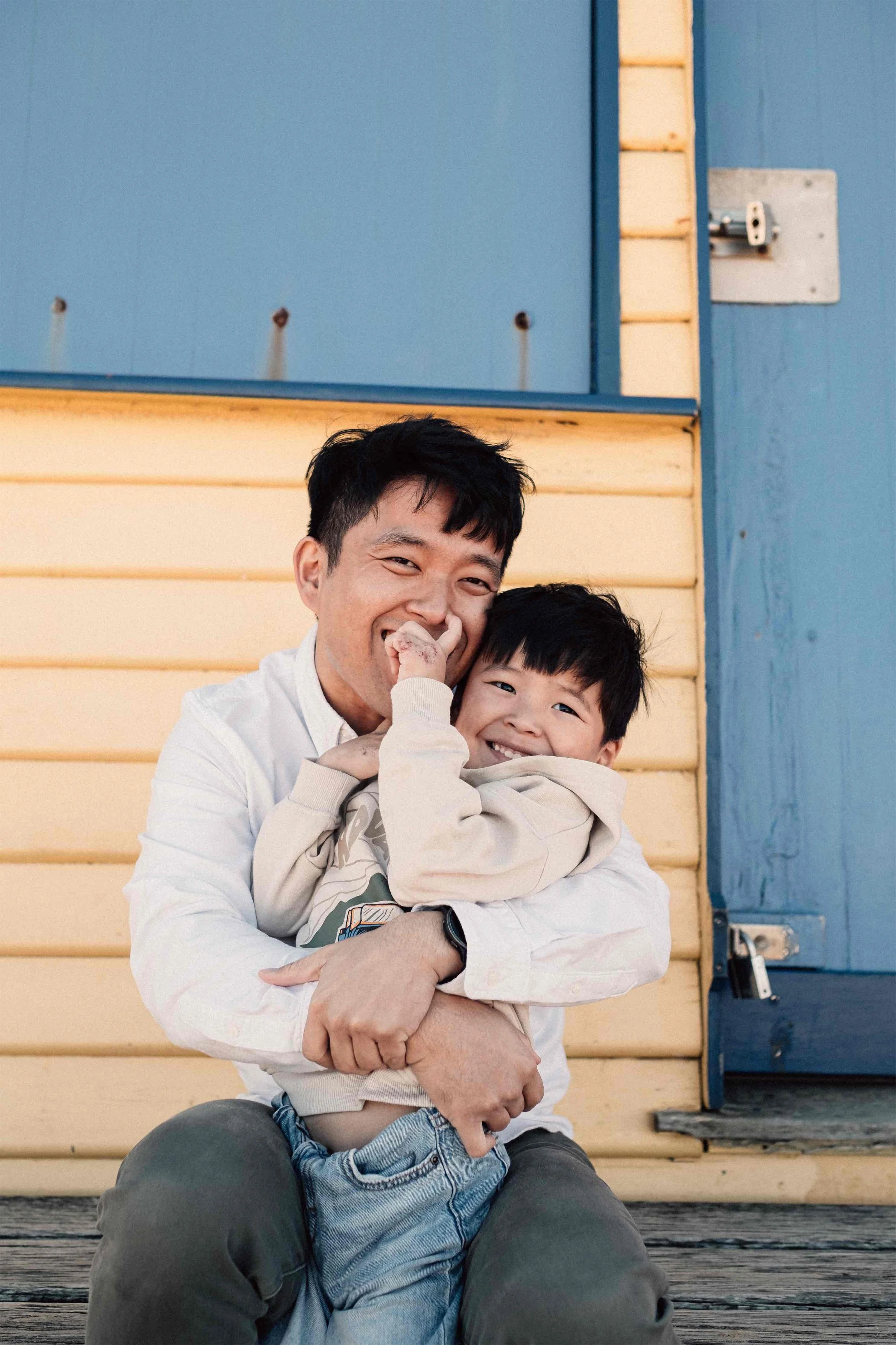 A man and a young boy sitting on a wooden bench, smiling and embracing each other; background features a colorful pastel blue and yellow building wall with a door. The man is wearing a white shirt and the boy is dressed in a light-colored hoodie and 