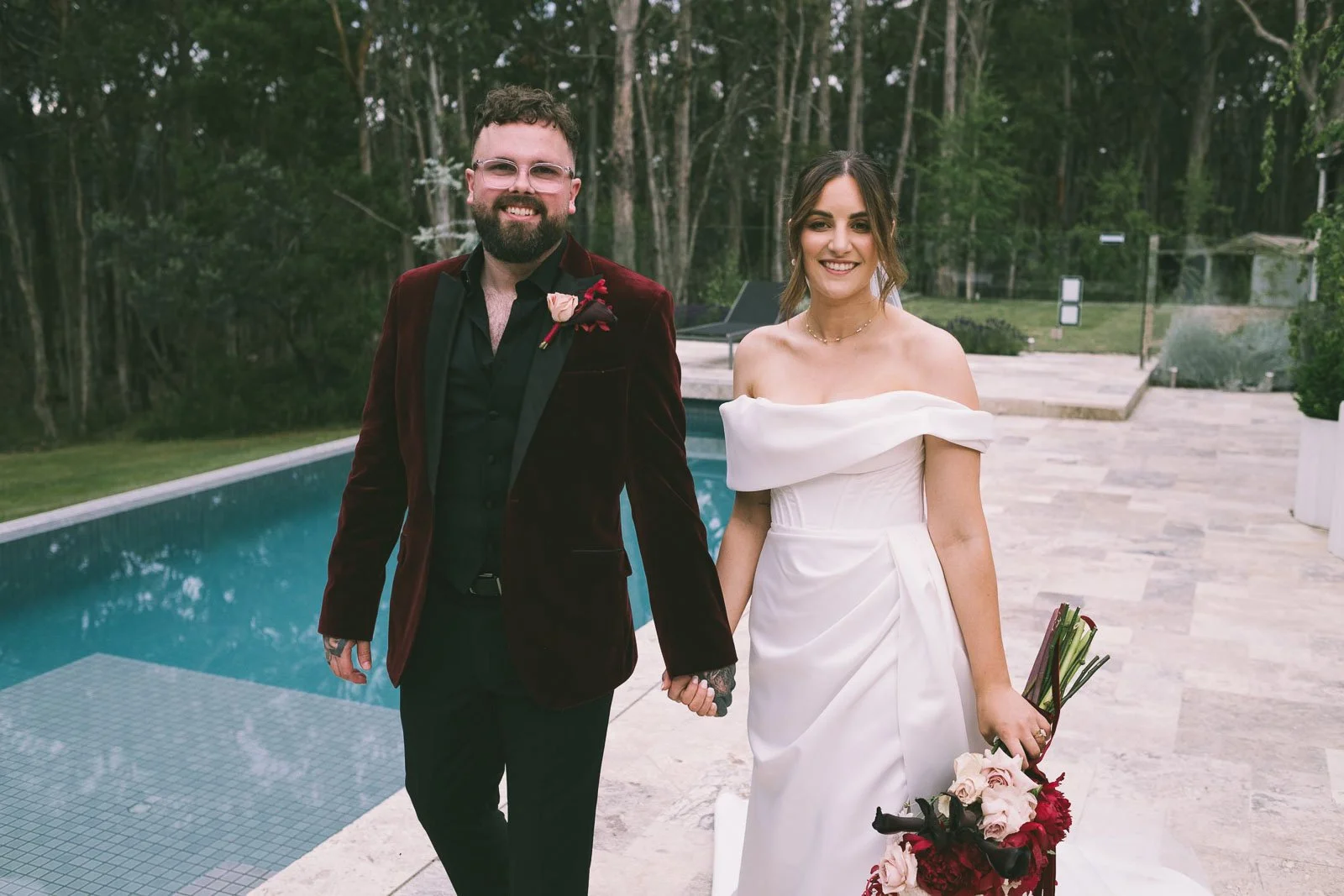 A bride and groom holding hands outdoors by a pool, smiling, with trees in the background.