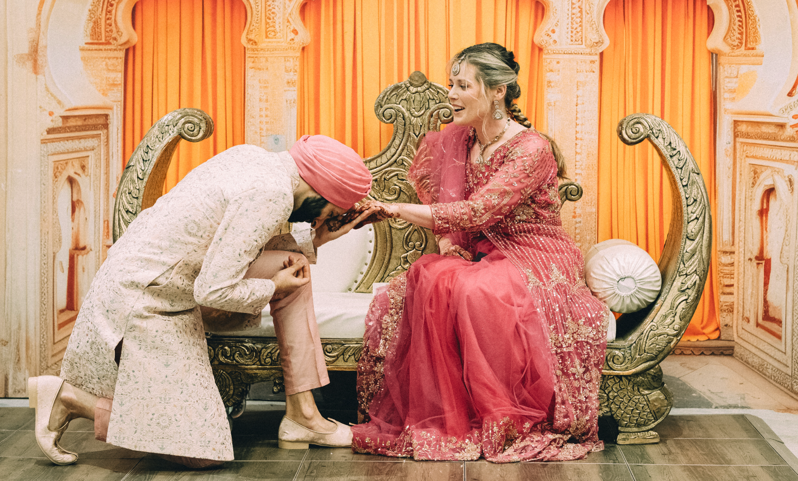 A woman dressed in a pink embroidered saree sitting on a decorative seat, touching foreheads with a man in traditional Indian attire, who is kneeling and bowing his head.