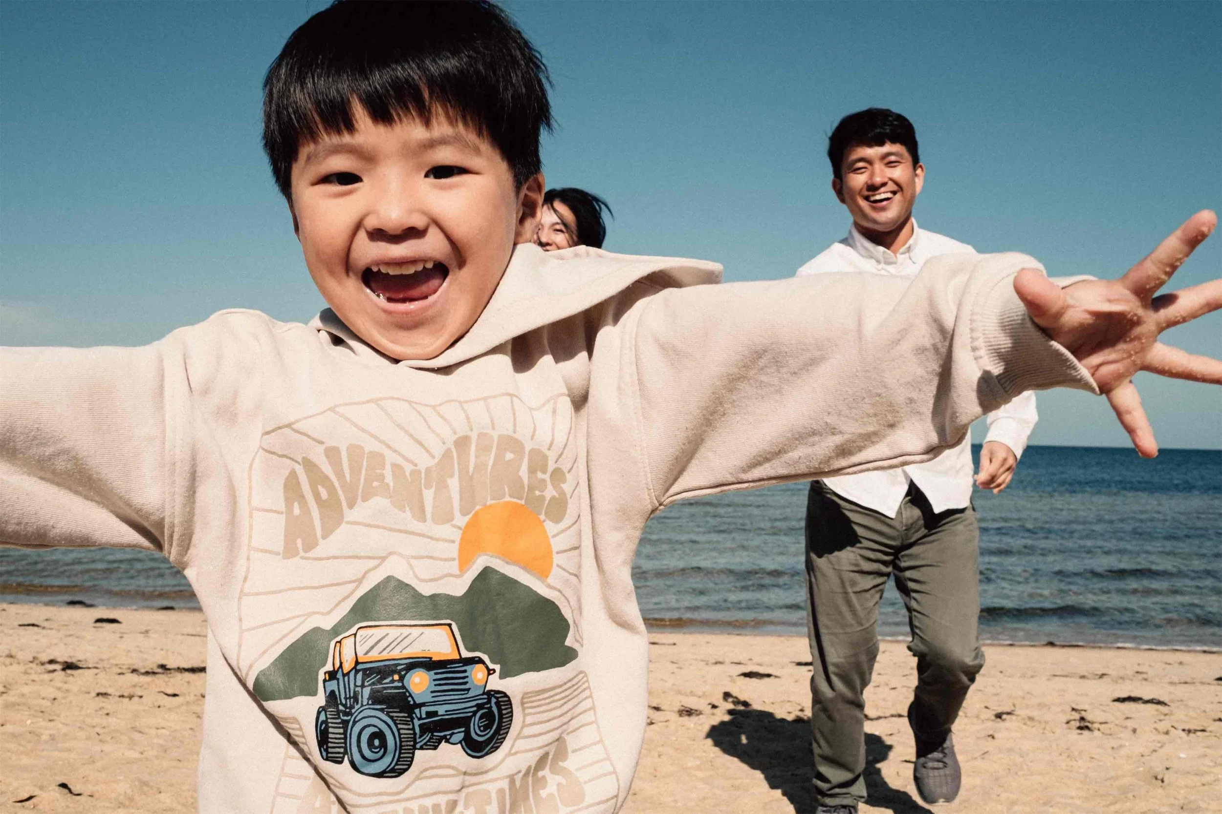 Three people, including a young boy in the foreground, are smiling and running on a sandy beach near the water, with a clear blue sky above.