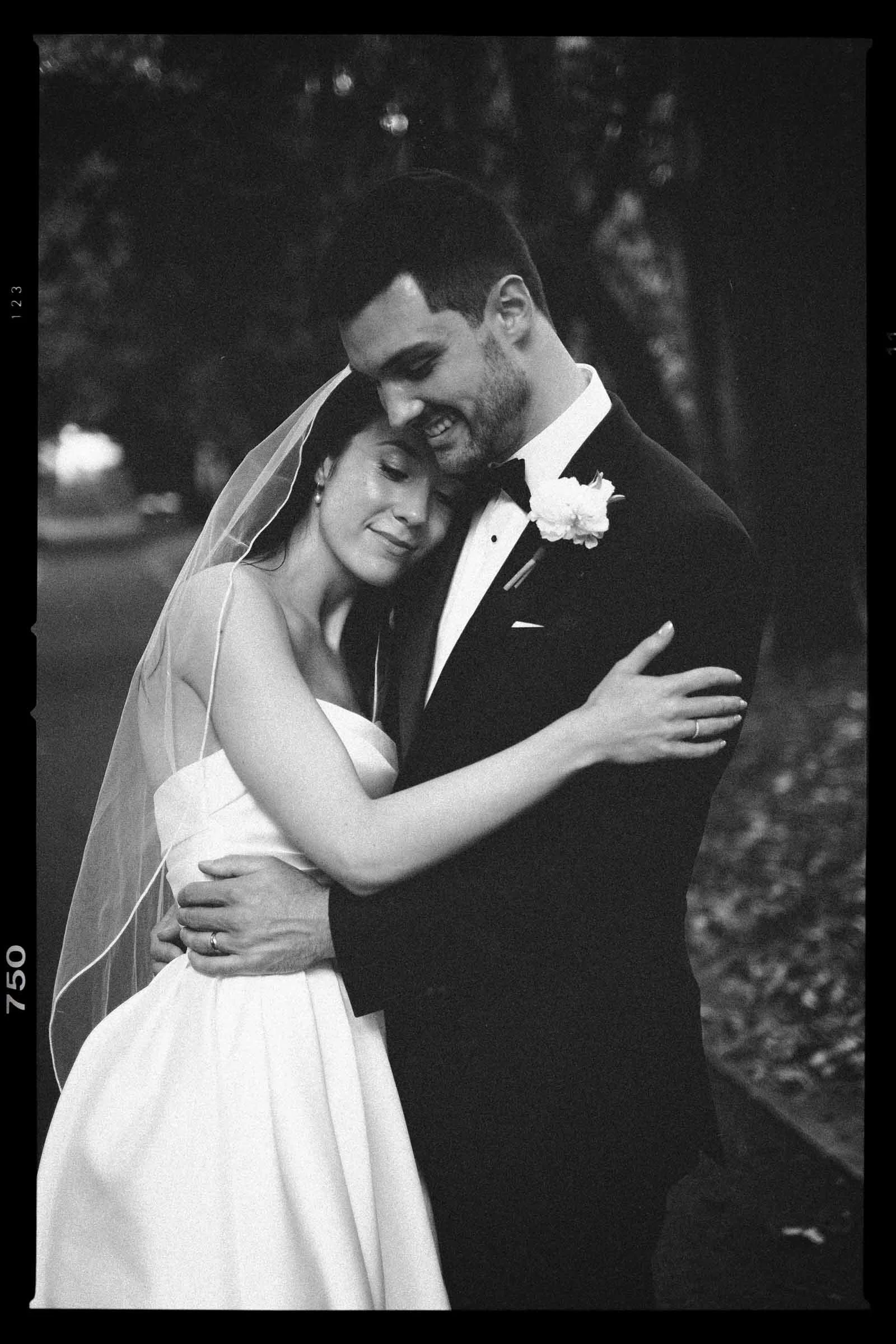 A black and white photo of a bride and groom embracing outdoors, smiling, with trees in the background. The bride is wearing a strapless wedding dress and veil, and the groom is in a tuxedo with a boutonnière.