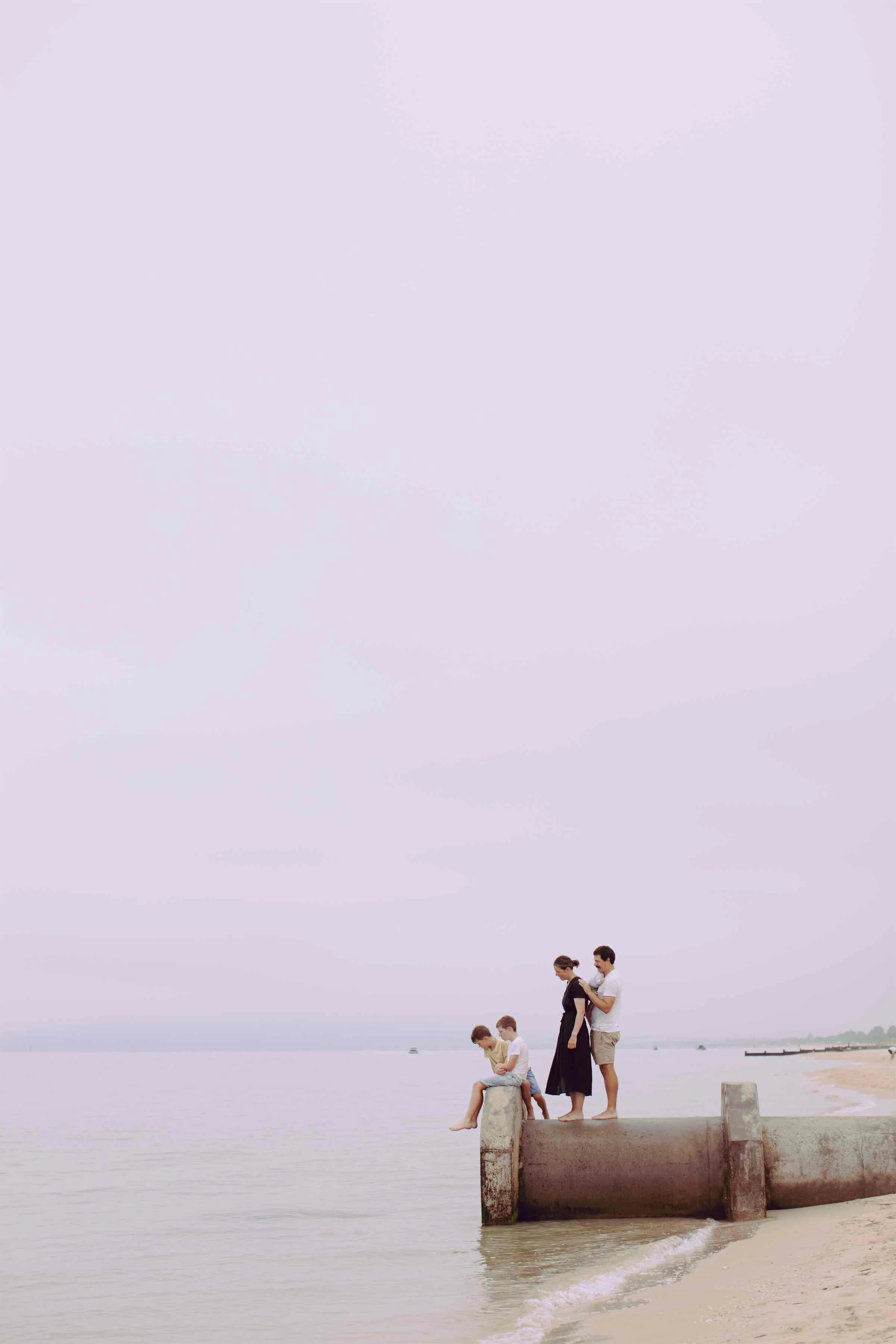 A family of five, including two adults and three children, is standing on a concrete pier by the water at the beach. The sky is overcast, and the family appears to be relaxing and enjoying the view of the sea.