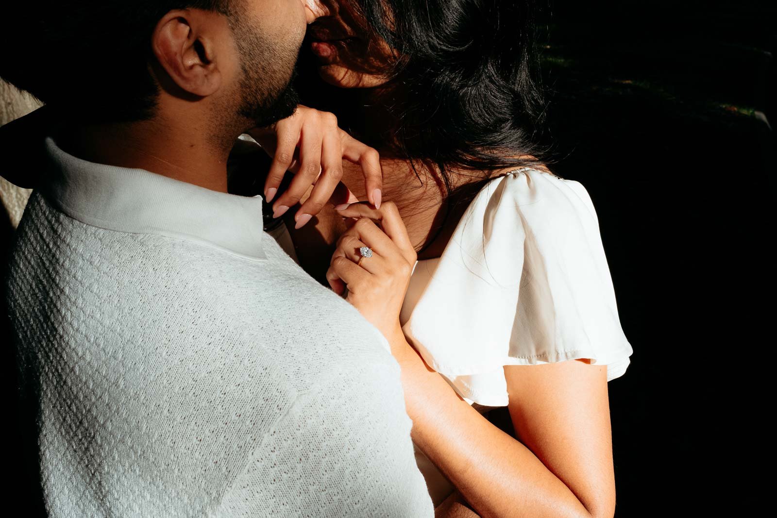 A man and woman are shown close together, with the woman wearing a white dress and an engagement ring, touching the man's shoulder and the man holding her hand.
