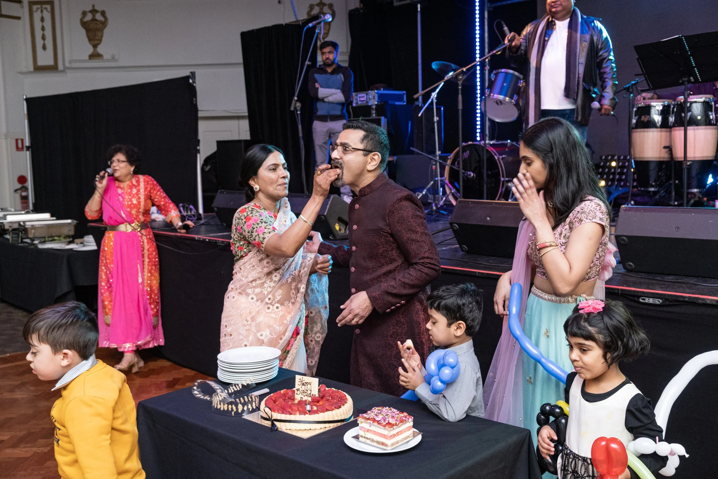People celebrating birthday with cakes, balloons, and decorations. A woman in a saree feeding a man in a traditional outfit, children in colorful clothes around the table, a woman with a microphone, and a band playing on stage in the background.