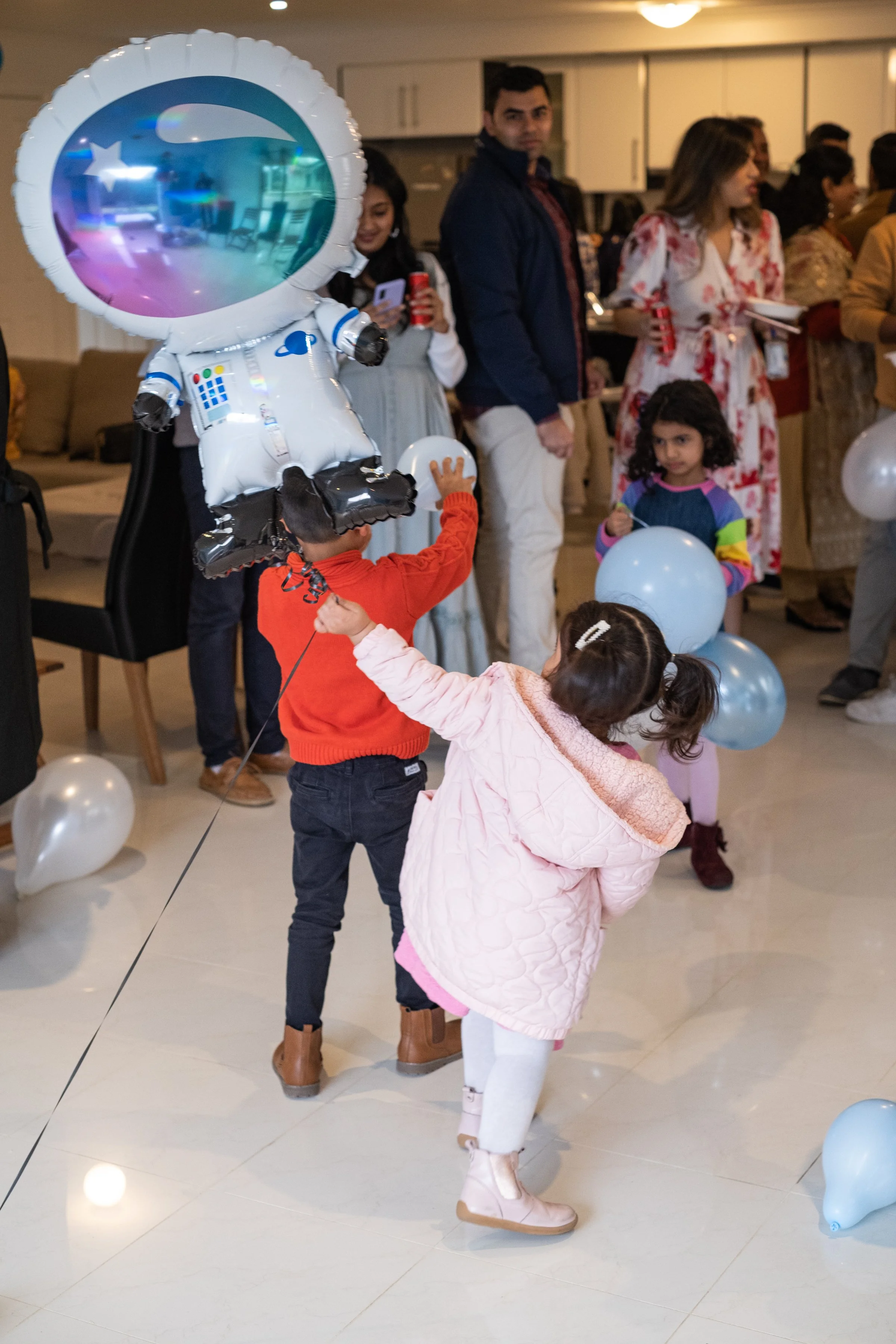 Children and adults celebrating at a party with balloons, including a large astronaut-shaped balloon, in a well-lit indoor space.