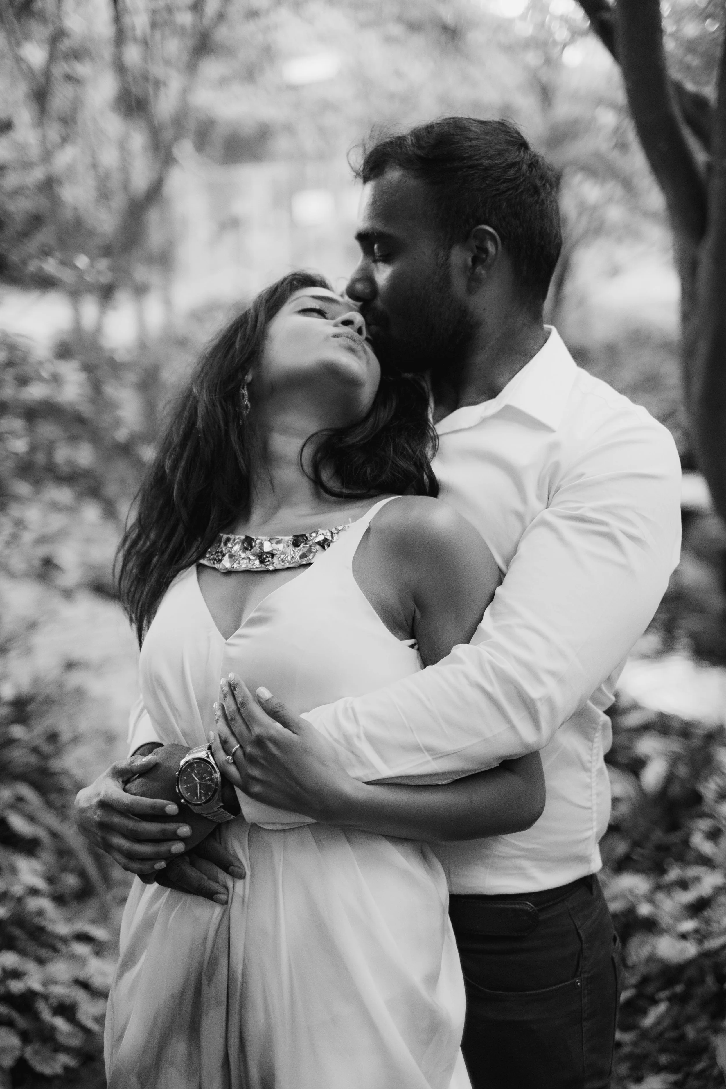 A black-and-white photo of a man and woman embracing each other outdoors among trees. The man is kissing the woman's forehead as she closes her eyes. They are holding each other tightly, and the woman's dress has decorative embellishments around the 