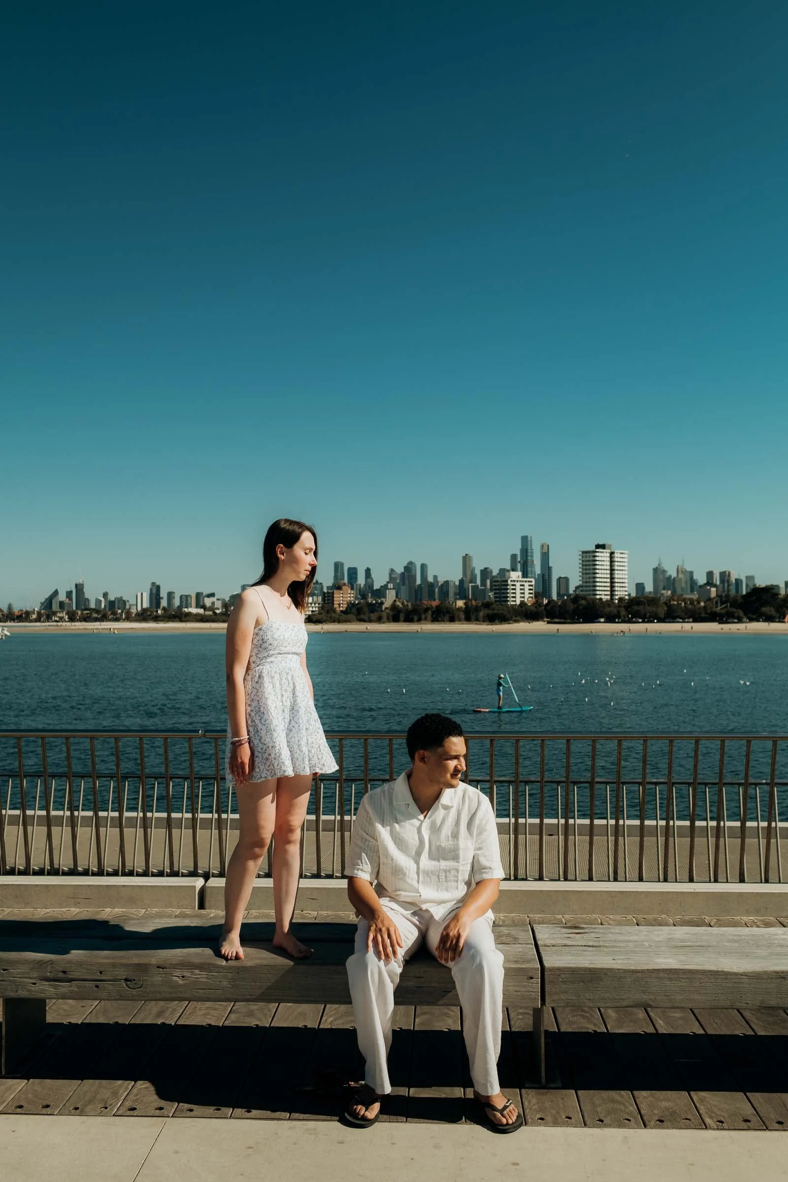 A woman standing on a deck, looking toward the water with a city skyline in the background, and a man sitting on the deck near her, facing forward.